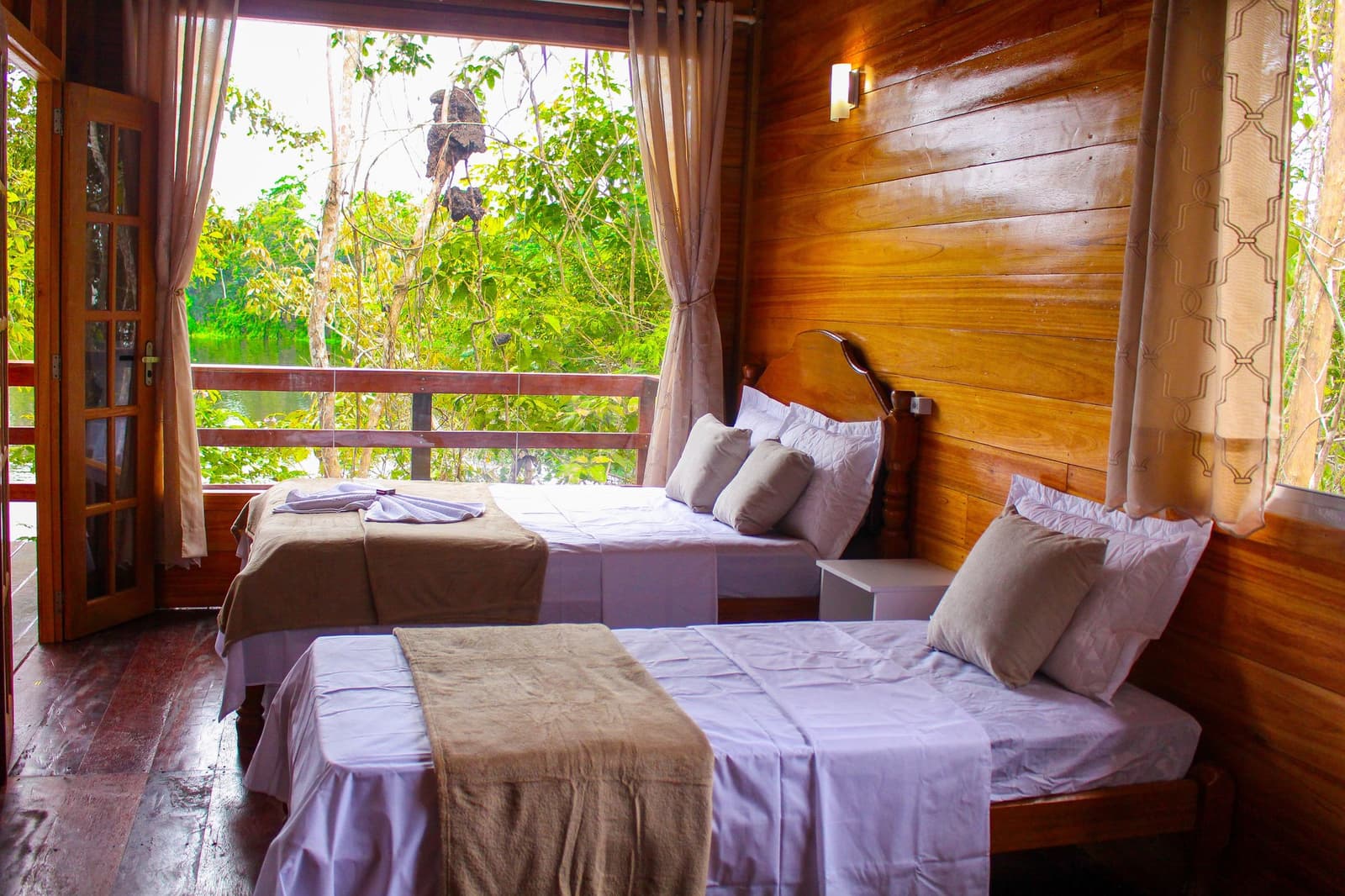 Frische Bettwäsche, warmes Holz und der Regenwald direkt vor dem Balkon. Zweibettzimmer mit weißer Bettwäsche und warmen Holzwänden in einer Amazonas-Dschungellodge, mit einem Balkon, der auf einen grünen Wasserweg mit Regenwaldvegetation blickt