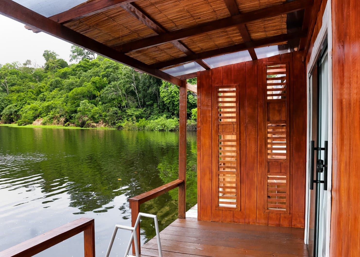 Schweres Holz, stilles Wasser, Dschungel auf drei Seiten. Holzdeck und Veranda mit Strohdach einer Öko-Lodge im brasilianischen Amazonas, mit Holzlamellenfenstern zum ruhigen Schwarzwasserfluss und dichtem Regenwald dahinter