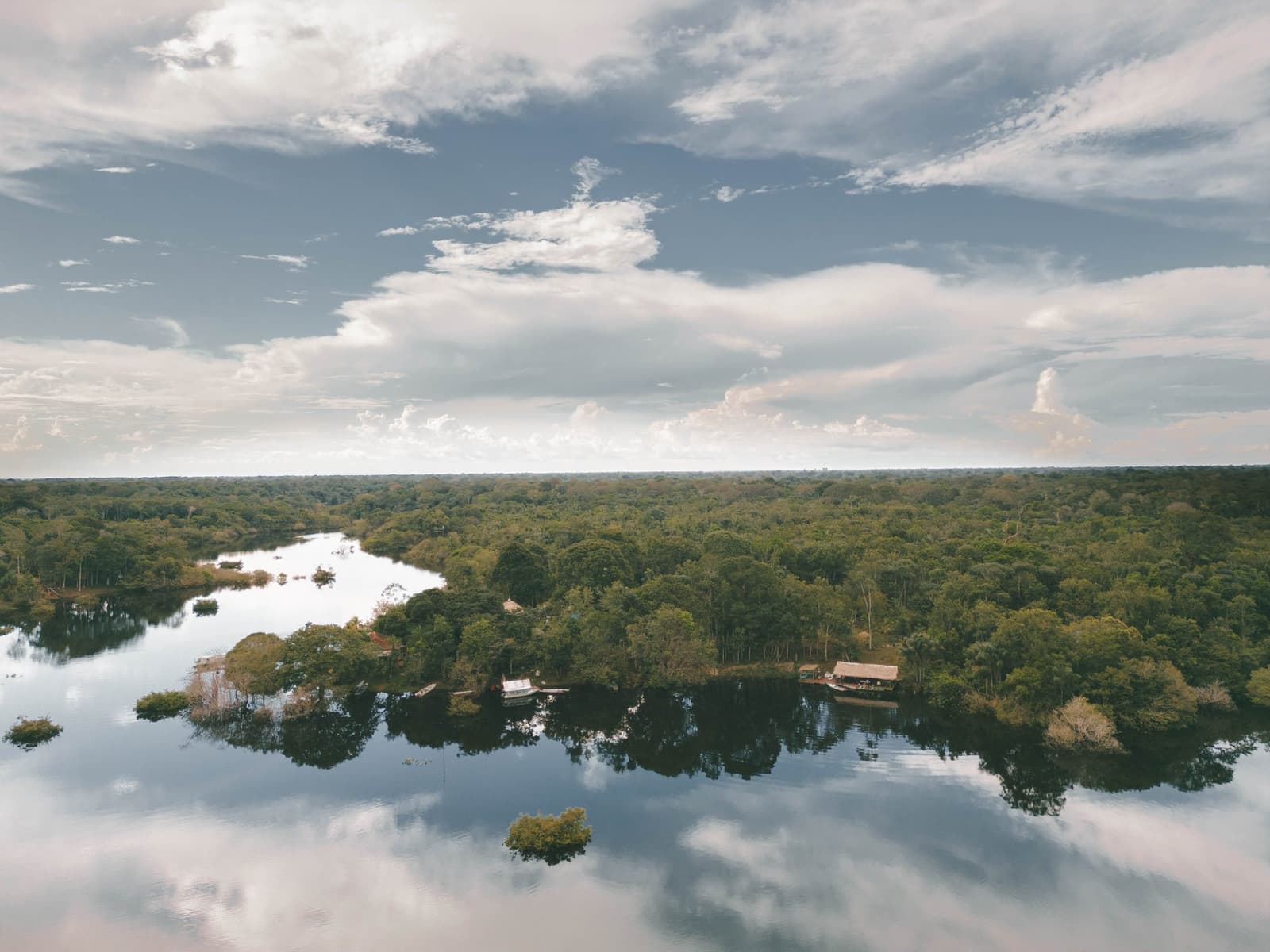 Endloser Baldachin, die Lodge kaum sichtbar am Flussufer. Luftaufnahme eines ruhigen Schwarzwasserflusses, der sich durch dichten tropischen Regenwald schlängelt, mit einem Reetdach-Lodge und einem kleinen Boot am bewaldeten Ufer