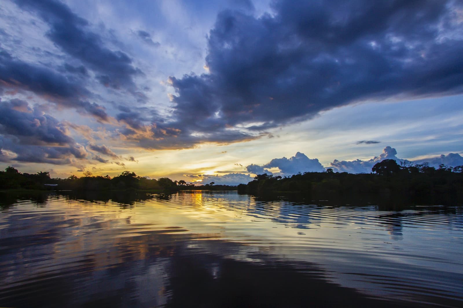 Gewitterwolken, goldenes Wasser, Dschungelstille — Abend am Amazonas. Photo: David Nascimento @ Dramatischer Sonnenuntergang über einem breiten amazonischen Fluss beim Brazilian Amazon Jungle Tour, mit goldenem Licht, das sich im stillen Wasser spiegelt, und silhouettierten Regenwaldwipfeln unter tiefblauen Gewitterwolken