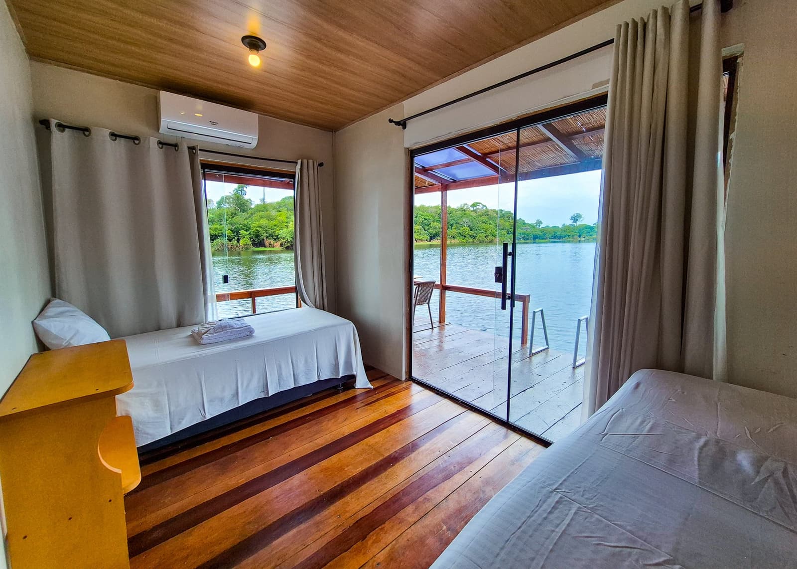 Einschlafen mit Flussrauschen und Regenwald direkt vor der Tür. Zweibettzimmer einer Amazonas-Lodge mit Holzboden, Klimaanlage und einer Glasschiebetür, die auf eine überdachte Veranda mit Blick auf einen ruhigen Fluss und dichten Regenwald führt