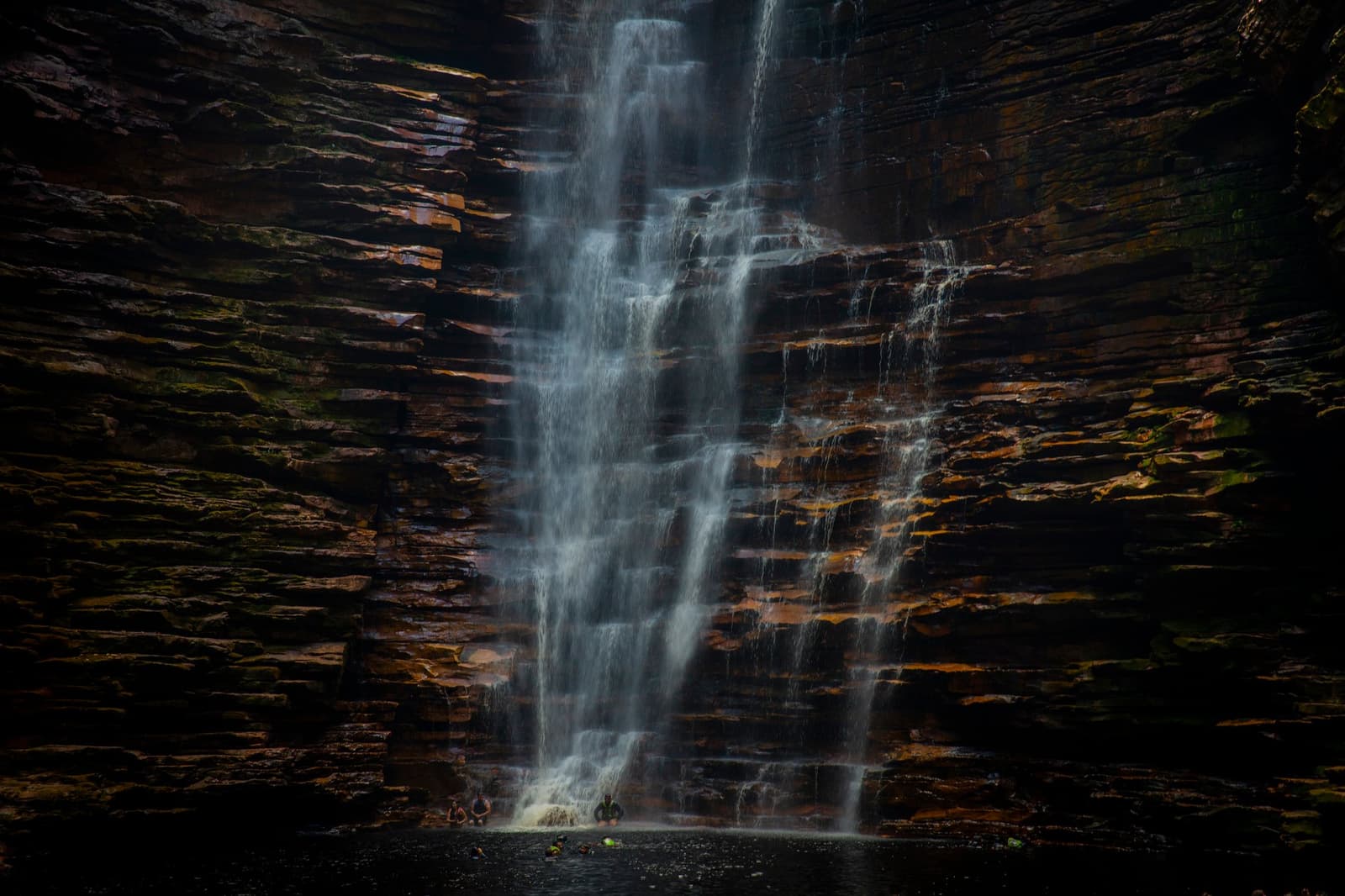 Água fria, pedra antiga e uma sensação de que você merecia isso. Cachoeira de múltiplos degraus caindo sobre paredões de arenito em camadas durante uma trilha na Chapada Diamantina, com duas figuras minúsculas visíveis na base da piscina natural