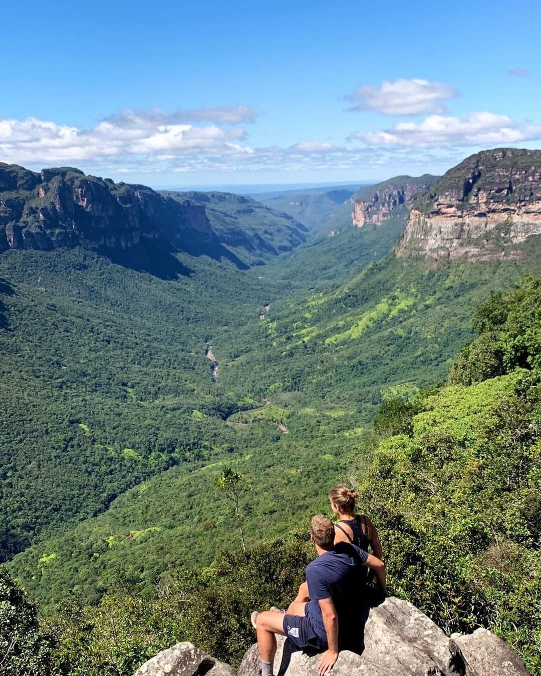 O vale fala por si, e eles só precisam ouvir. Dois caminhantes sentados numa pedra contemplam um imenso cânion coberto de vegetação durante uma trilha na Chapada Diamantina, com paredes de arenito e um rio serpenteando lá embaixo