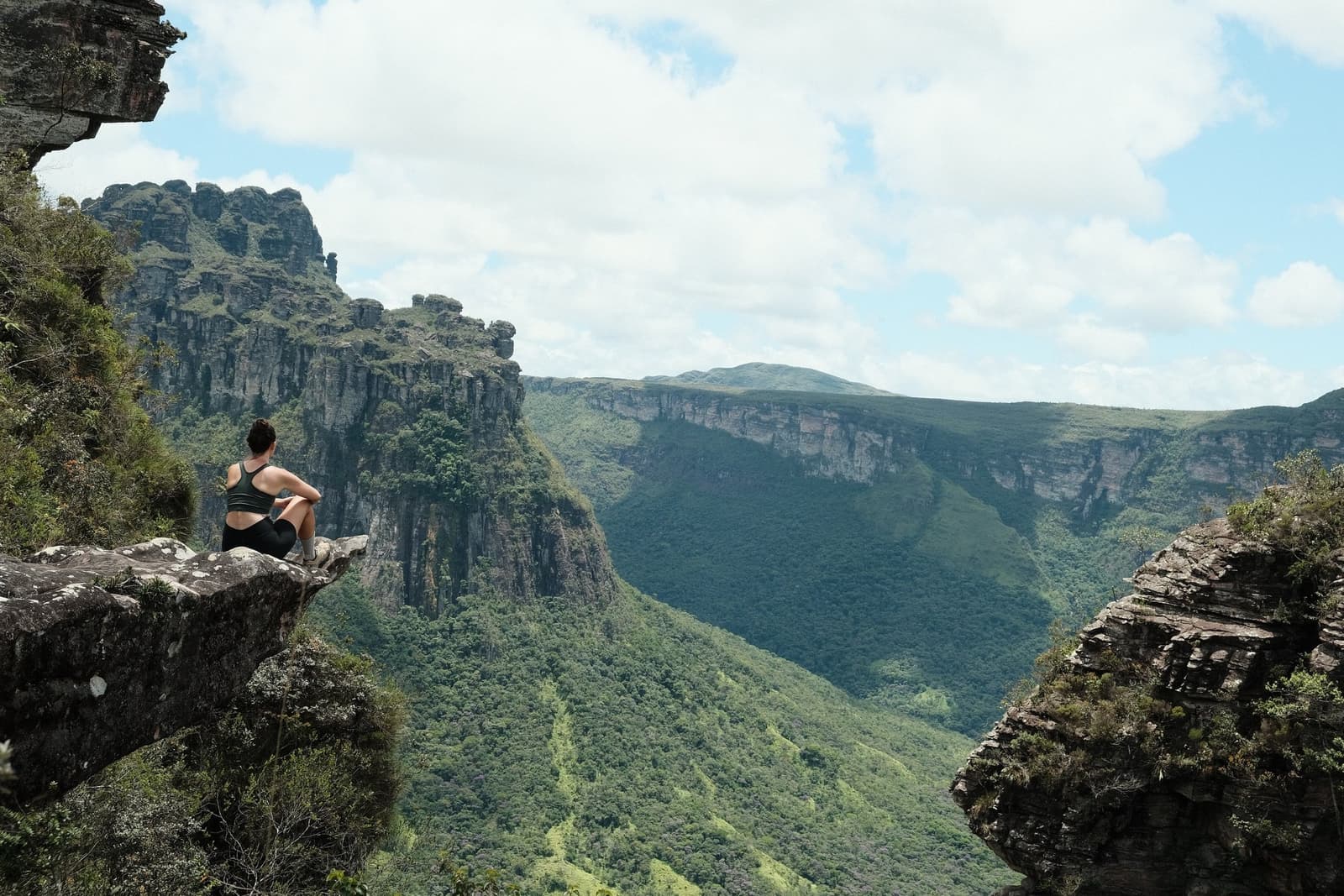 Tem vista que a gente guarda pra sempre, mesmo sem tirar foto. Uma caminhante sentada em uma saliência rochosa observa um vasto canyon verde com falésias de arenito durante uma trilha na Chapada Diamantina