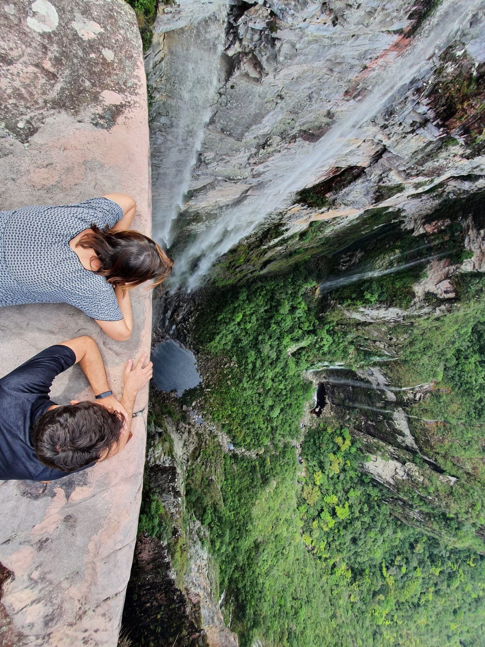 Na Chapada, a vista mais honesta é a que dá medo de olhar. Dois caminhantes deitados sobre uma saliência de rocha nua observam diretamente o fundo de uma falésia íngreme, onde uma fina cachoeira percorre as paredes do canyon cobertas de musgo lá embaixo