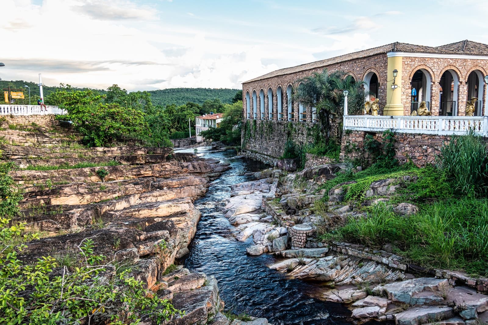Rio e pedra na mesma conversa de sempre, bem no coração da cidade. Um casarão histórico de pedra com arcadas se debruça sobre um canal rochoso cortado no arenito estratificado em uma cidade de partida para a trilha na Chapada Diamantina, com morros cobertos de mata ao fundo