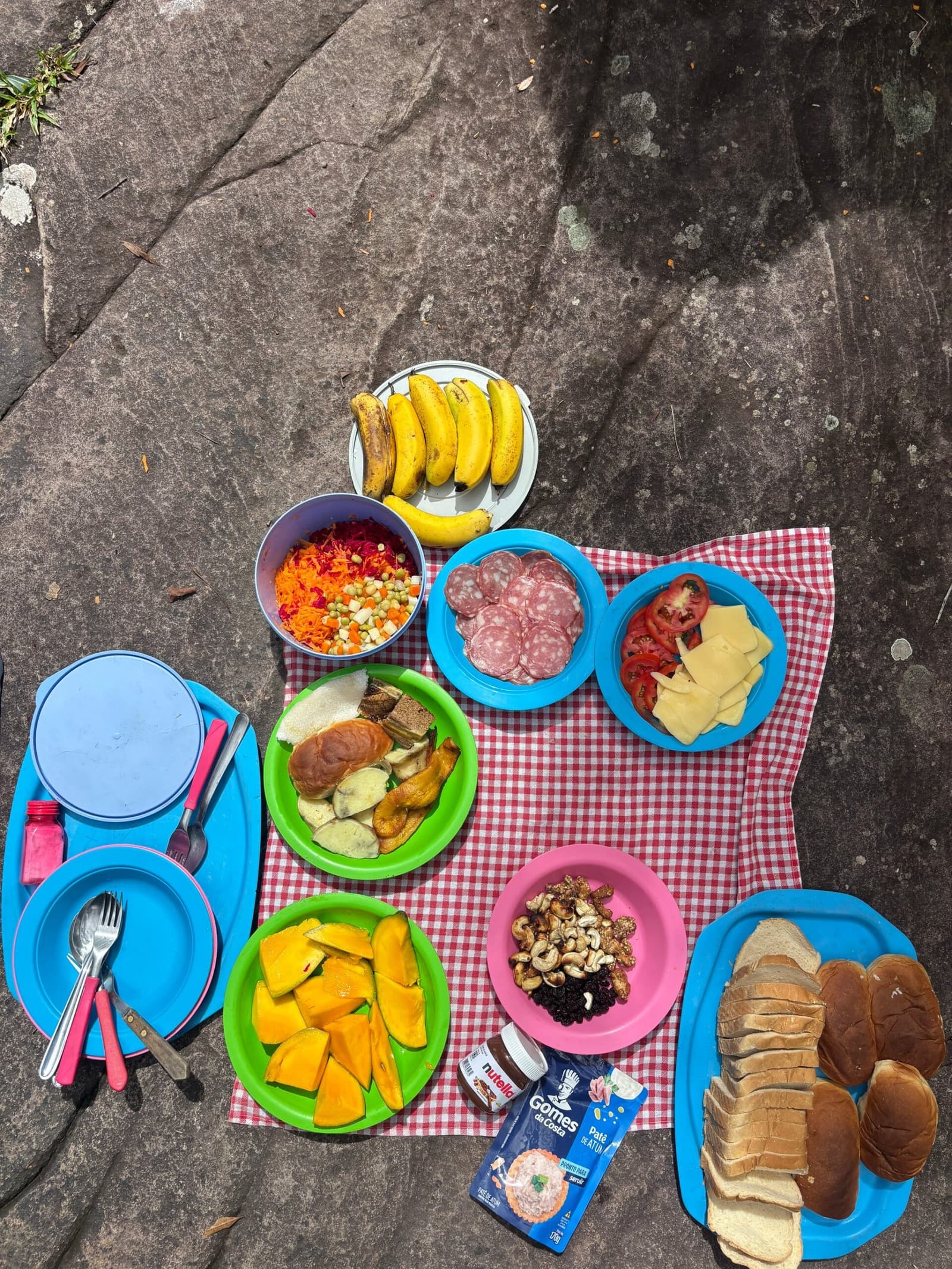 Almoço no meio da Chapada: simples, farto e muito bem-vindo. Vista aérea de um piquenique colorido sobre uma rocha plana durante uma trilha na Chapada Diamantina, com pratos de frutas, salame, queijo, tomates, legumes ralados, castanhas de caju, passas, bananas, sanduíches e pão de forma sobre uma toalha xadrez vermelha