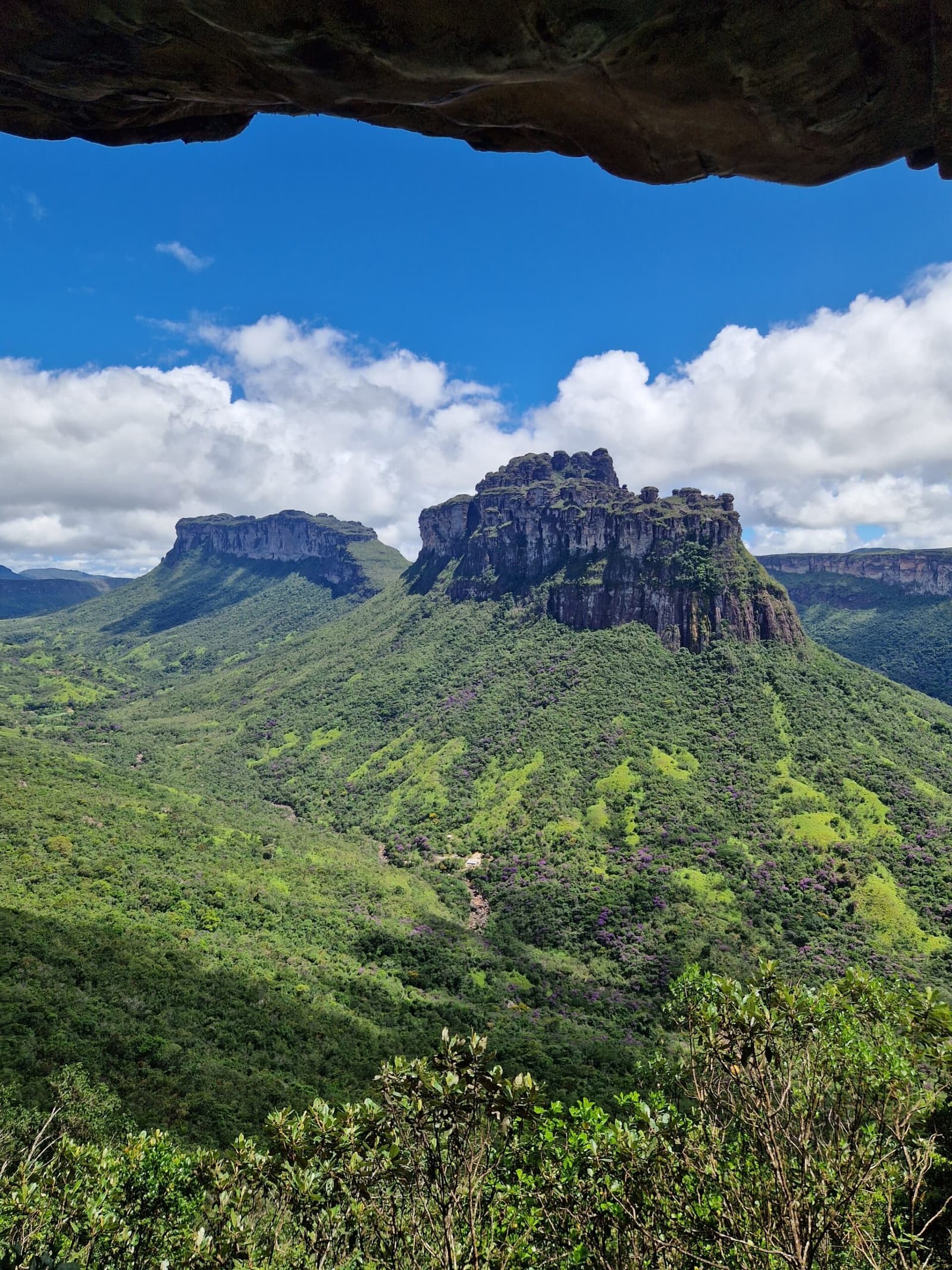 A pedra fez a moldura. O resto é trilha. Um paredão rochoso emoldura a vista de um morro de topo plano erguendo-se acima de colinas cobertas de vegetação exuberante em uma trilha na Chapada Diamantina, sob um céu azul intenso com nuvens brancas