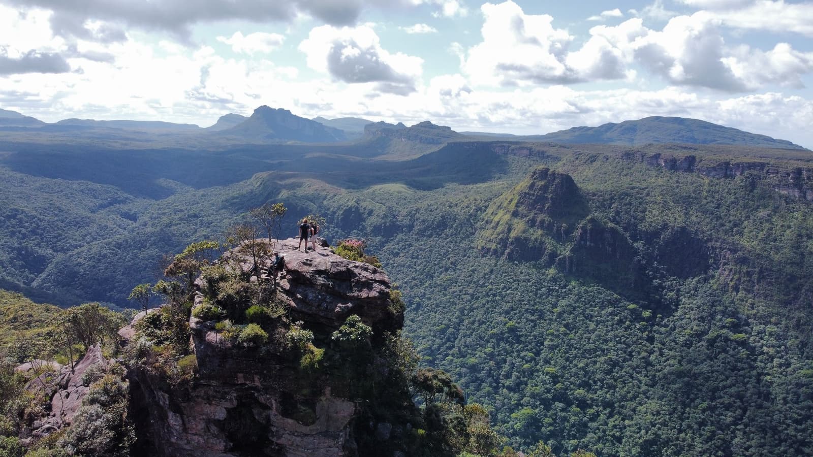 Chegar aqui a pé tem um sabor que nenhuma foto consegue carregar sozinha. Dois caminhantes em cima de um afloramento de arenito com musgo durante uma trilha na Chapada Diamantina, com um imenso canyon coberto de mata e cadeias de montanhas se estendendo até o horizonte sob céu parcialmente nublado