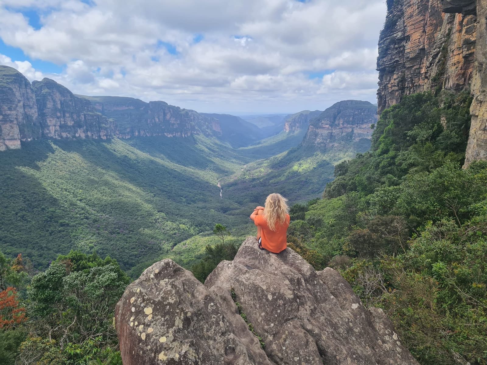 Chegar aqui com as próprias pernas muda completamente a forma de ver o vale. Uma caminhante de jaqueta laranja sentada sobre uma rocha de granito contempla um vale verde e amplo ladeado por paredões de arenito durante uma trilha na Chapada Diamantina
