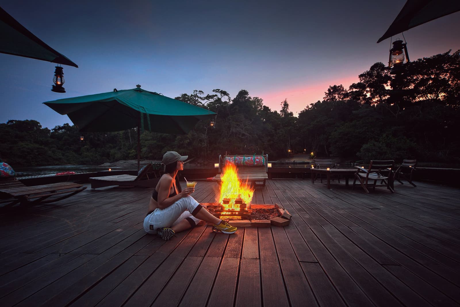Feuer, Dschungel, ein kühles Getränk, der Abend braucht nichts mehr. Eine Gästin sitzt neben einem offenen Feuer auf der Holzterrasse der Cristalino Jungle Lodge, hält einen Cocktail in der Hand, während ein rosa-blauer Abendhimmel über dem Regenwalddach leuchtet und hängende Laternen die Szene erhellen
