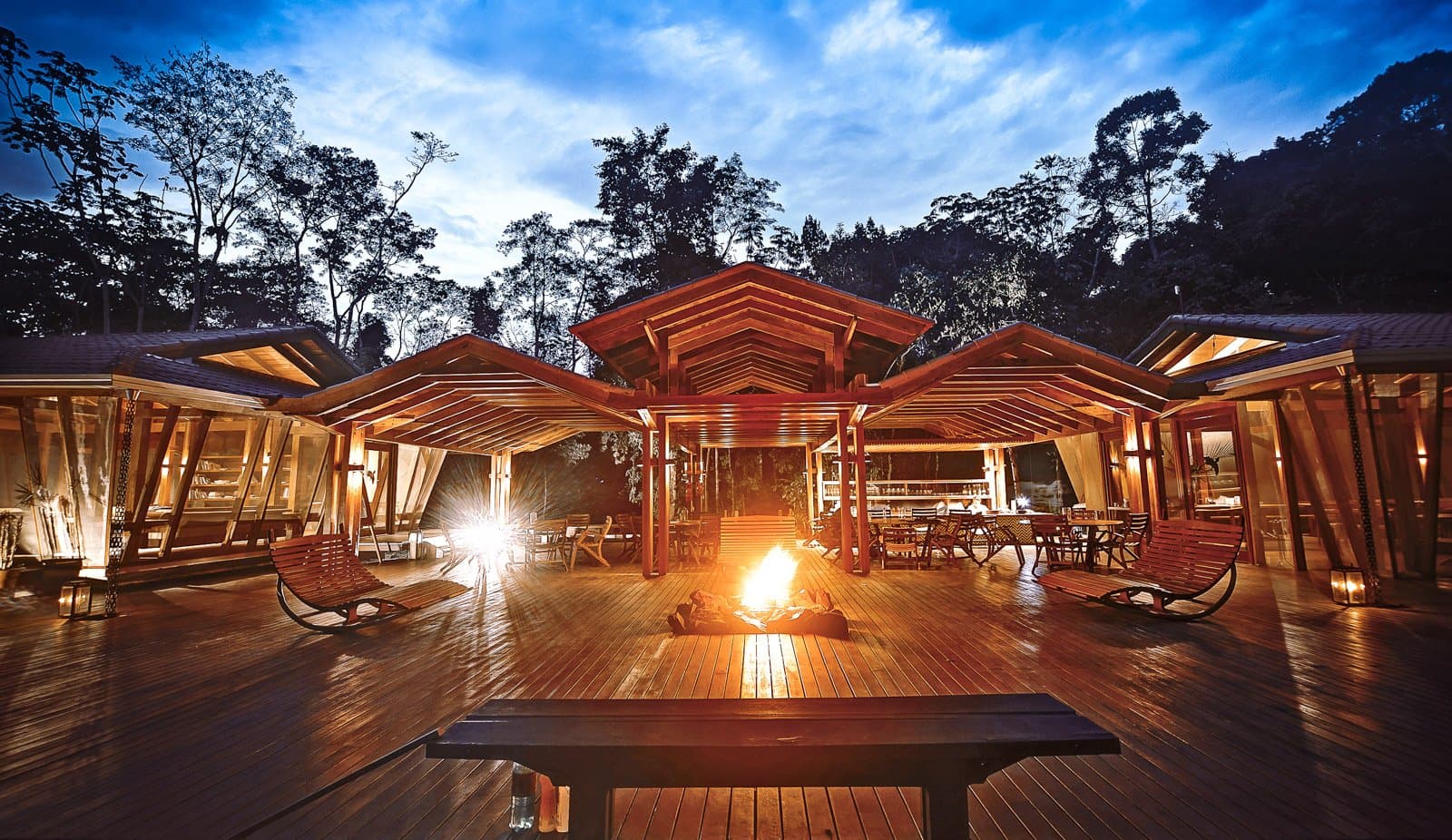 Wenn der Regenwald schweigt, übernimmt das Feuer das Gespräch. Die offene Holzterrasse des Cristalino Jungle Lodge leuchtet in der Abenddämmerung: Ein knisterndes Lagerfeuer brennt in der Mitte, Schaukelstühle flankieren die Seiten, eine Bar ist im Hintergrund zu sehen und hohe Regenwaldsbäume zeichnen sich gegen den tiefblauen Abendhimmel ab