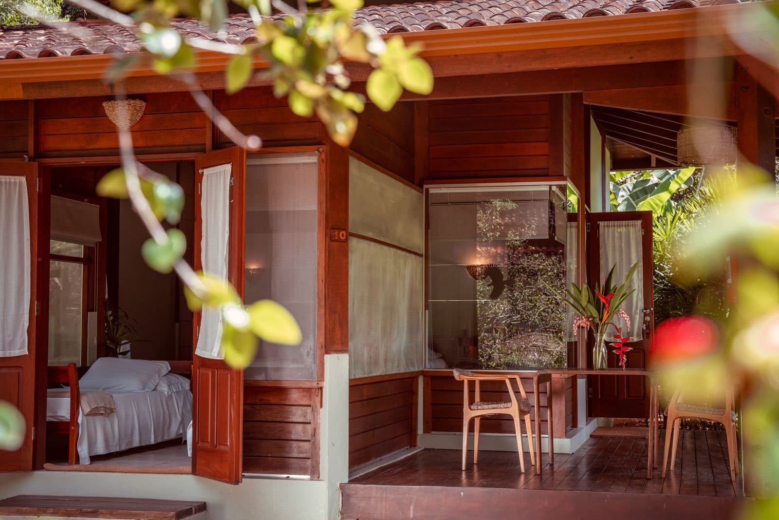 Türen auf, Regenwald rein. So beginnt der Morgen hier. Holzbungalow im Cristalino Jungle Lodge mit offenen Türen, sichtbarem Bett mit weißer Bettwäsche, überdachter Veranda mit Holzstühlen und tropischen Blütenpflanzen