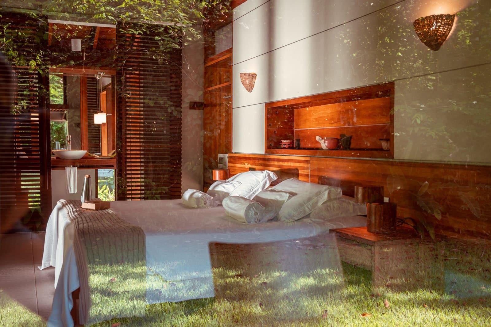 Schlafen, während der Regenwald sanft durch die Wände schaut. Blick durch eine Glasscheibe in ein Bungalow-Schlafzimmer des Cristalino Jungle Lodge mit Doppelbett in weißer Bettwäsche, warmem Holzkopfteil mit eingebauten Regalen, geflochtenen Wandleuchten und einem Badezimmer mit Holzlamellen-Fensterläden