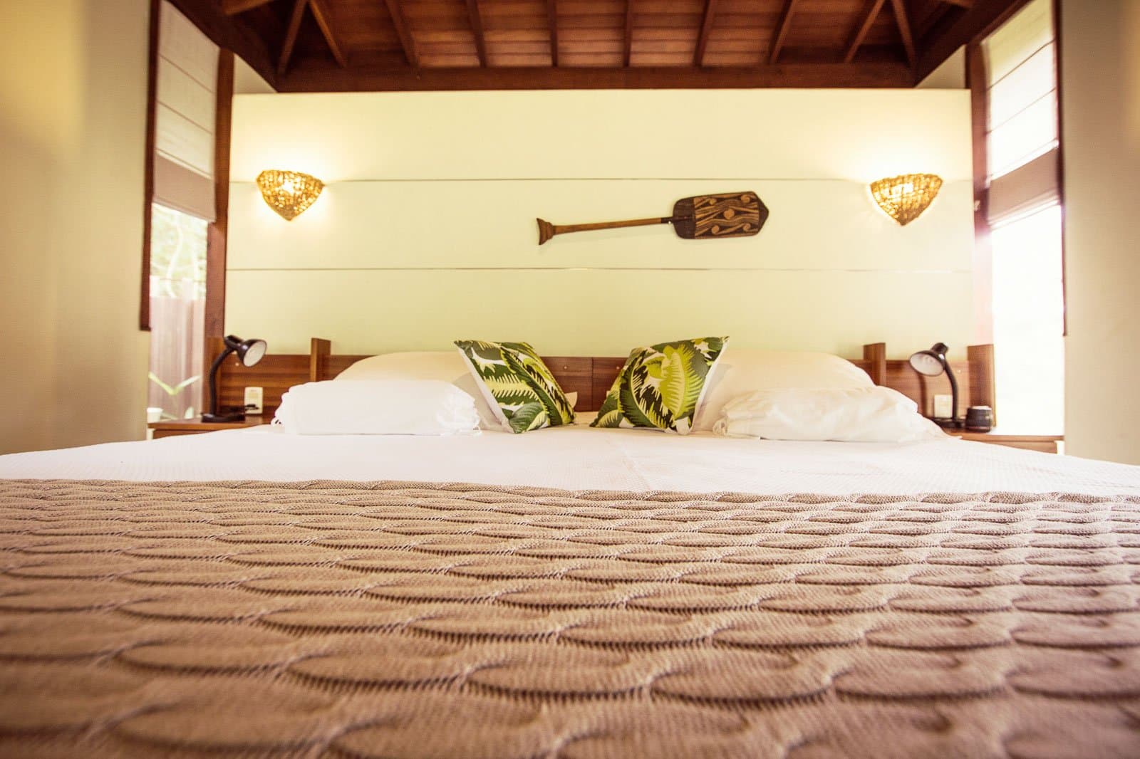 Frische Bettwäsche, ein Holzpaddel an der Wand und der Regenwald gleich vor dem Fenster. Kingsize-Bett mit tropischen Blattmuster-Kissen, Holzkopfteil, geflochtenen Wandleuchten und einem geschnitzten Paddel an der Wand in einem Bungalow der Cristalino Jungle Lodge