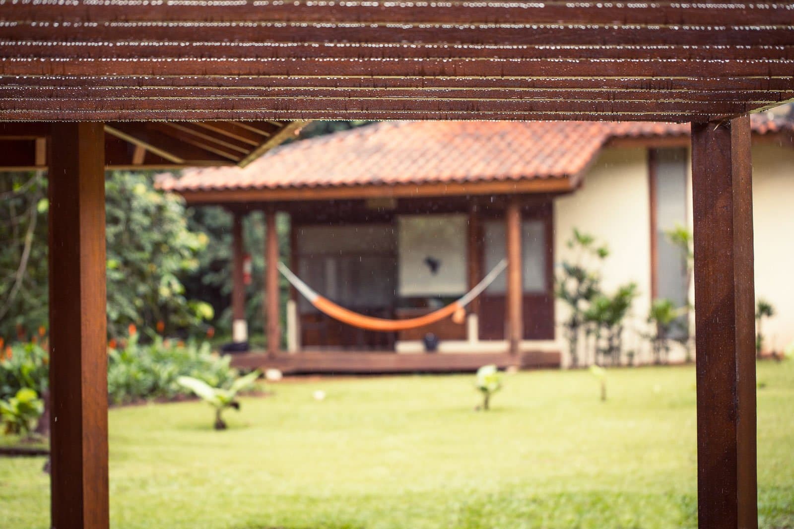 Regen auf dem Dach, eine Hängematte in Reichweite, mehr braucht ein guter Nachmittag nicht. Regen fällt auf eine Holzpergola, die den Blick auf ein Bungalow des Cristalino Jungle Lodge mit einer orangefarbenen Hängematte auf der Veranda und sattgrünem Rasen rahmt