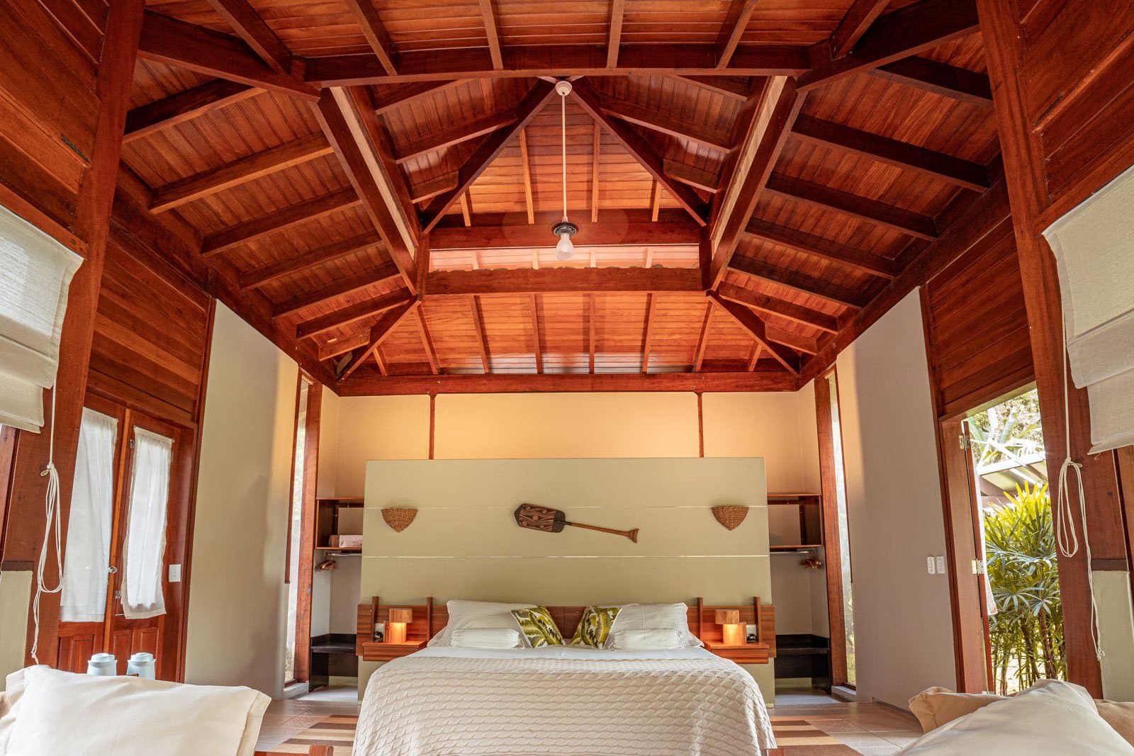 Schlafen unter Hartholzbalken, das Paddel an der Wand verrät, dass der Fluss ganz nah ist. Geräumiges Bungalow-Interieur im Cristalino Jungle Lodge mit hoch gewölbter Hartholzdecke, Kingsize-Bett mit weißer Bettwäsche und blattgemusterten Kissen, warmem Kerzenlicht auf den Nachttischen und einem dekorativen Holzpaddel über dem Kopfteil