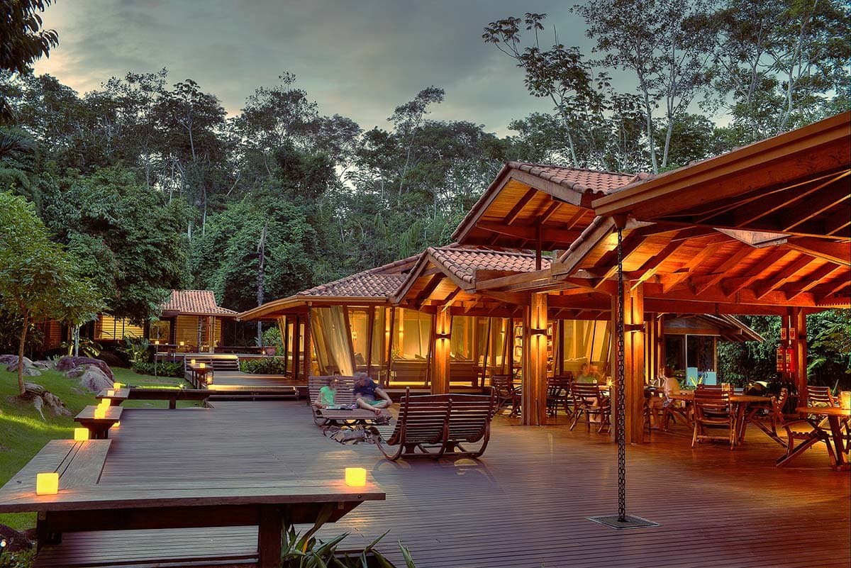 Die Abenddämmerung gehört hier der Lodge, nicht dem Lärm. Die Holzterrasse der Cristalino Jungle Lodge leuchtet in der Abenddämmerung im Schein von Kerzen und warmen Lampen, während Gäste auf Schaukelstühlen entspannen und dichter Regenwald im Hintergrund aufragt