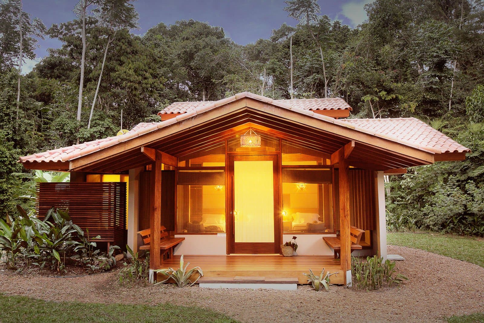 Hier schläft man gut, der Regenwald summt das Schlaflied. Ein holzgerahmter Bungalow im Cristalino Jungle Lodge erstrahlt in warmem Bernsteinlicht in der Abenddämmerung, mit Terrakottadach und Holzterrasse vor dichtem Regenwalddach