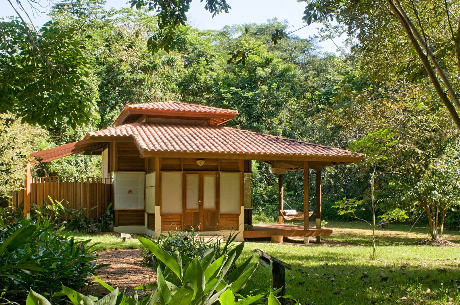 Eigene vier Wände im Regenwald, die Hängematte macht das Ankommen leicht. Ein Holzbungalow mit terrakottafarbenem Ziegeldach und einer überdachten Holzterrasse mit Hängematte, eingebettet in üppigen Tropenwald im Cristalino Jungle Lodge