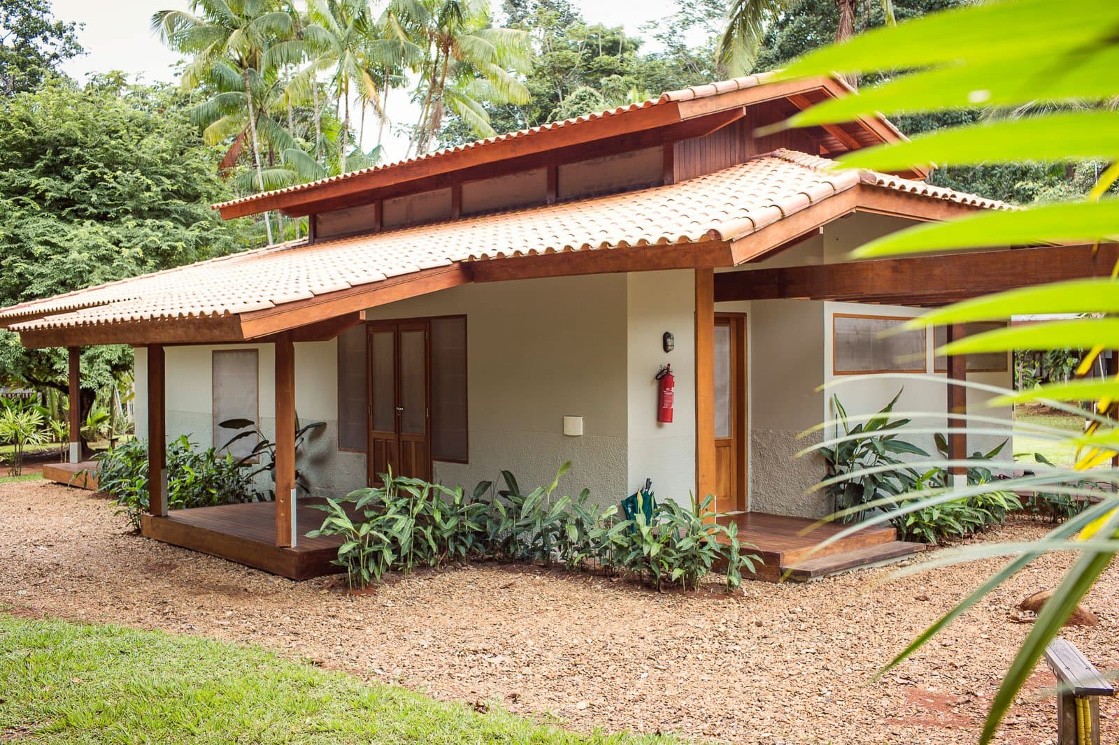 Genug Komfort, um den Dschungel draußen kurz zu vergessen, aber er wartet geduldig. Außenansicht eines Chalets der Cristalino Jungle Lodge mit Terrakotta-Ziegeldach, Holzveranda und üppiger tropischer Bepflanzung vor einem dichten grünen Regenwaldhintergrund
