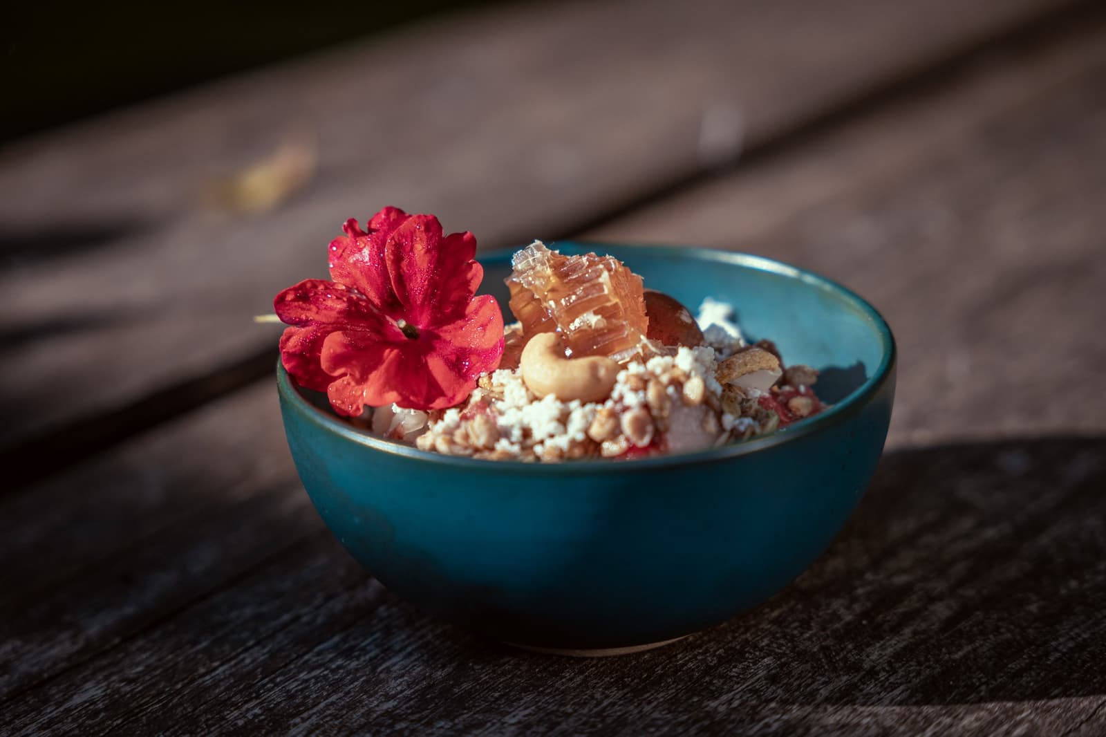 Frühstück im Regenwald, garniert mit einer Blüte statt einer Serviette. Eine blaugrüne Keramikschale im Cristalino Jungle Lodge, gefüllt mit Granola, Nüssen, Bienenwabe und frischen Früchten, garniert mit einer leuchtend roten essbaren Blüte auf einer verwitterten Holzoberfläche