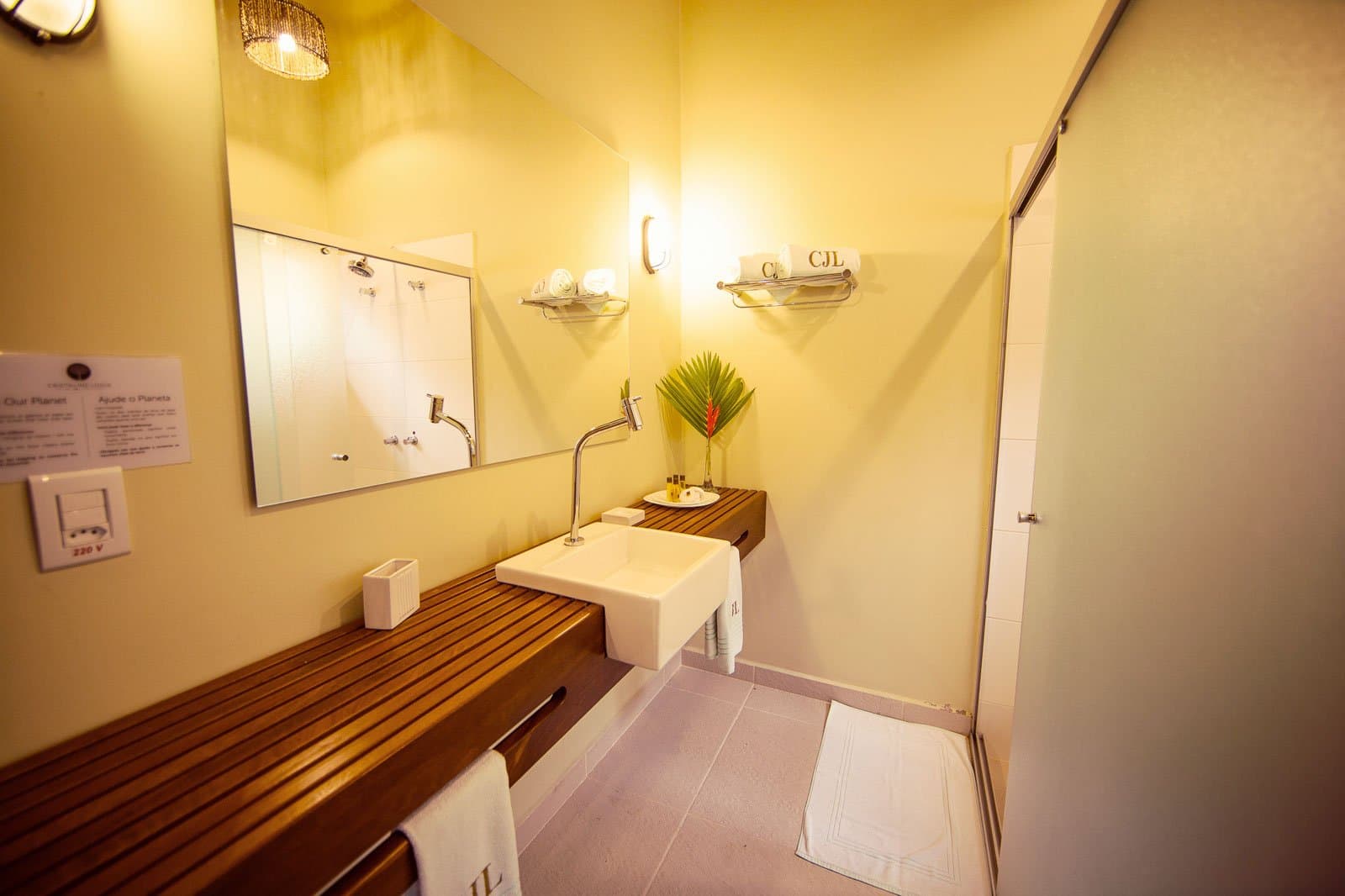 Mitten im Regenwald, und das Handtuch liegt trotzdem perfekt gefaltet bereit. Helles Badezimmer der Cristalino Jungle Lodge mit eckigem Aufsatzbecken auf einer Teakholz-Ablage, warmen Wandleuchten, monogrammierten Handtüchern und einer roten Heliconie neben der Dusche
