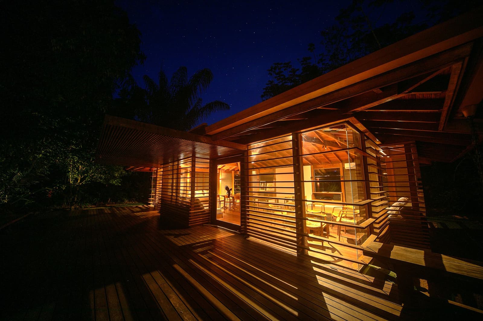 Abendessen im Regenwald, draußen Stille, drinnen ein warmes Willkommen. Das Cristalino Jungle Lodge erstrahlt nachts von innen, mit Holzlamellen-Wänden und einer breiten Holzterrasse in warmem Bernsteinlicht unter einem Sternenhimmel