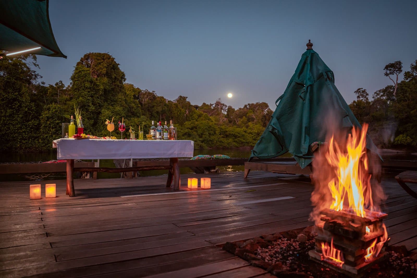 Der Abend gehört dem Feuer, den Drinks und einem Mond, der einfach nicht warten wollte. Eine offene Holzterrasse des Cristalino Jungle Lodge in der Abenddämmerung mit einem lodernden Lagerfeuer im Vordergrund, Papierlaternen auf dem Boden, einem festlich gedeckten Cocktailtisch mit bunten Getränken und einem Vollmond über dem Regenwalddach