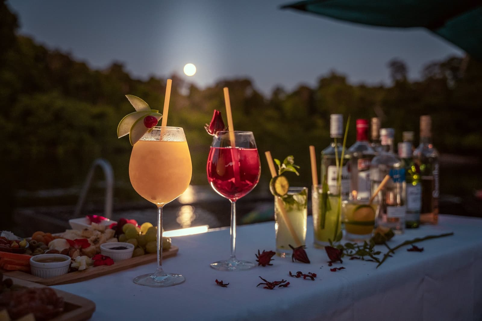 Vollmond, kühle Drinks, kein weiterer Plan nötig. Vier garnierte Cocktails und eine Käse-Charcuterie-Platte auf einem weißen Tisch im Cristalino Jungle Lodge, während ein Vollmond über dem Regenwalddach aufgeht