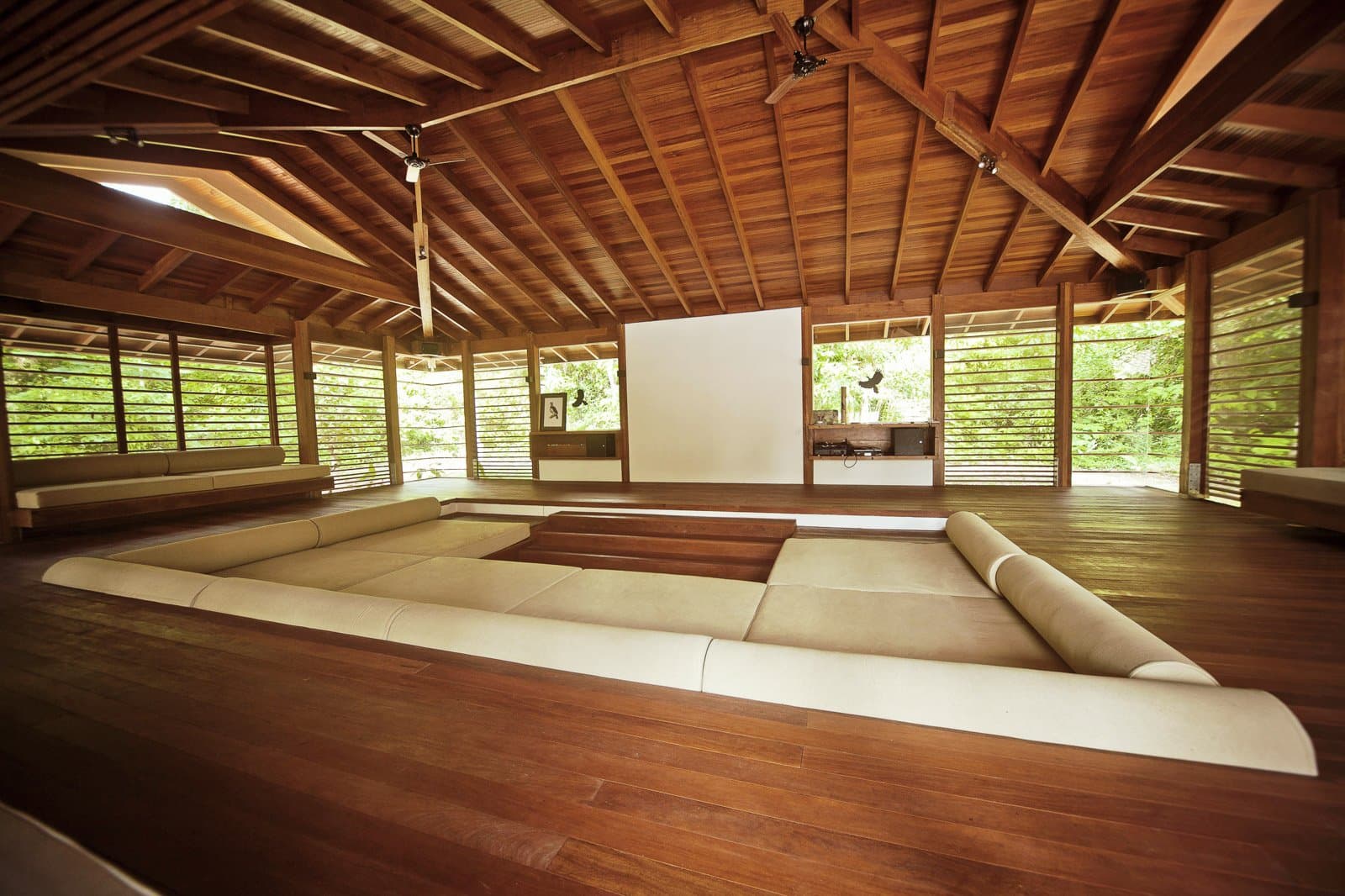 Hier sitzt man tief, atmet durch und hört dem Wald zu. Der offene Aufenthaltsraum der Cristalino Jungle Lodge mit versenktem Sitzbereich, cremefarbenen Kissen, poliertem Hartholzboden, einer großen Leinwand und einem gewölbten Holzdach mit Deckenventilatoren, umgeben von üppigem tropischem Grün.