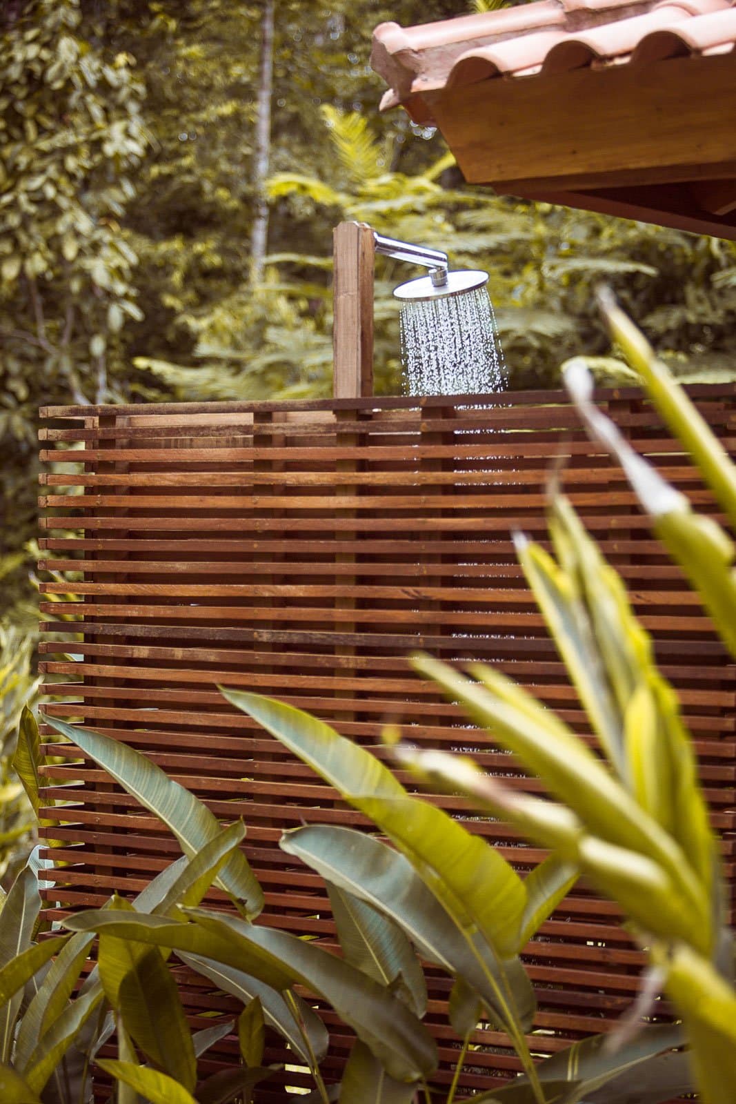 Abendliche Dusche, der Regenwald nur eine Armlänge entfernt. Ein Regenduschkopf im Freien mit fließendem Wasser über einer Sichtschutzwand aus Hartholzlamellen im Cristalino Jungle Lodge, eingerahmt von üppigem tropischem Blattwerk und einem Terrakottadach