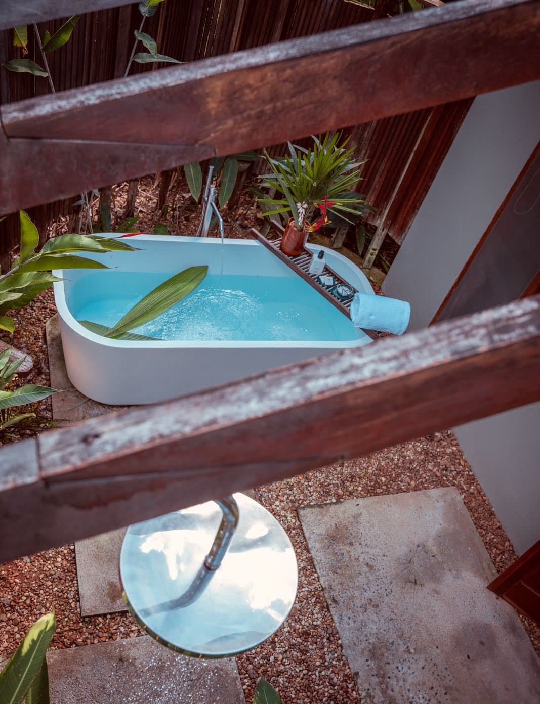 Das Beste am Regenwald? Manchmal einfach darin einweichen. Freistehende Outdoor-Badewanne mit klarem blauem Wasser im Cristalino Jungle Lodge, umgeben von tropischem Blattwerk, einem Holz-Badetablett mit Blumen und Pflegeprodukten sowie einer runden Außendusche, von oben durch ein rustikales Holzgeländer fotografiert