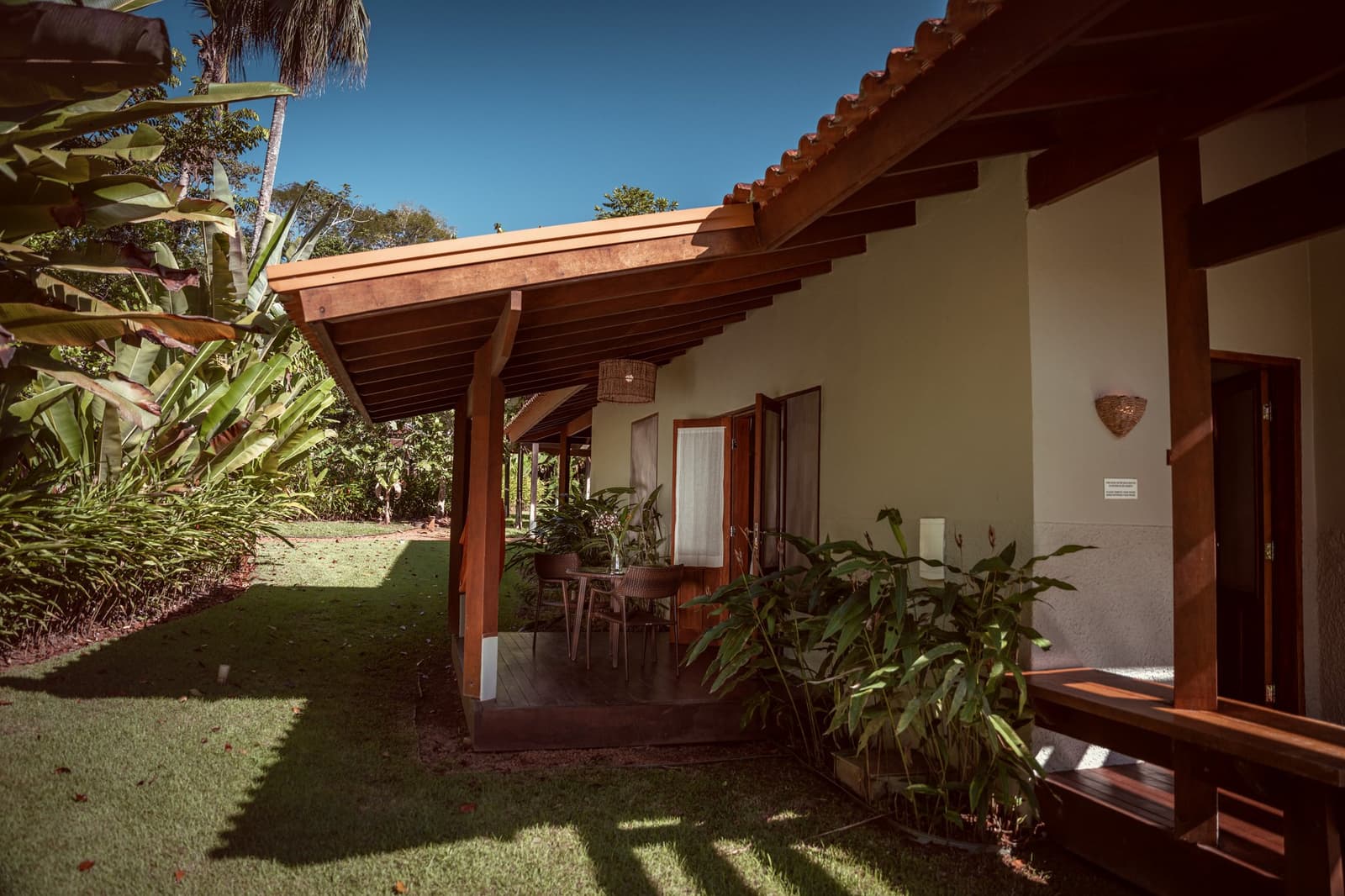 Ein eigenes Stück Grün, direkt vor der Tür. Überdachte Holzveranda der Cristalino Jungle Lodge mit einem kleinen Esstisch im Freien, Rattanlampe und üppigem tropischem Garten unter blauem Himmel