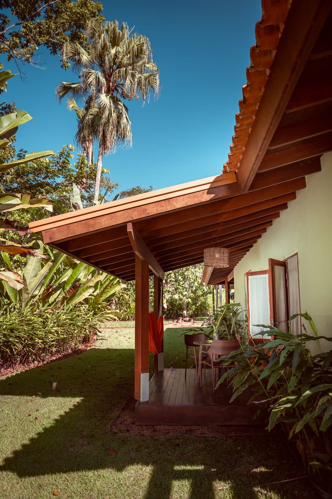 Schatten, Rattanlampe, Amazonas direkt hinter dem Rasen. Überdachte Hartholzveranda des Cristalino Jungle Lodge mit Rattanlampe, Rattanstühlen und üppigem tropischen Garten mit Bananenstauden und einer hohen Palme unter tiefblauem Himmel