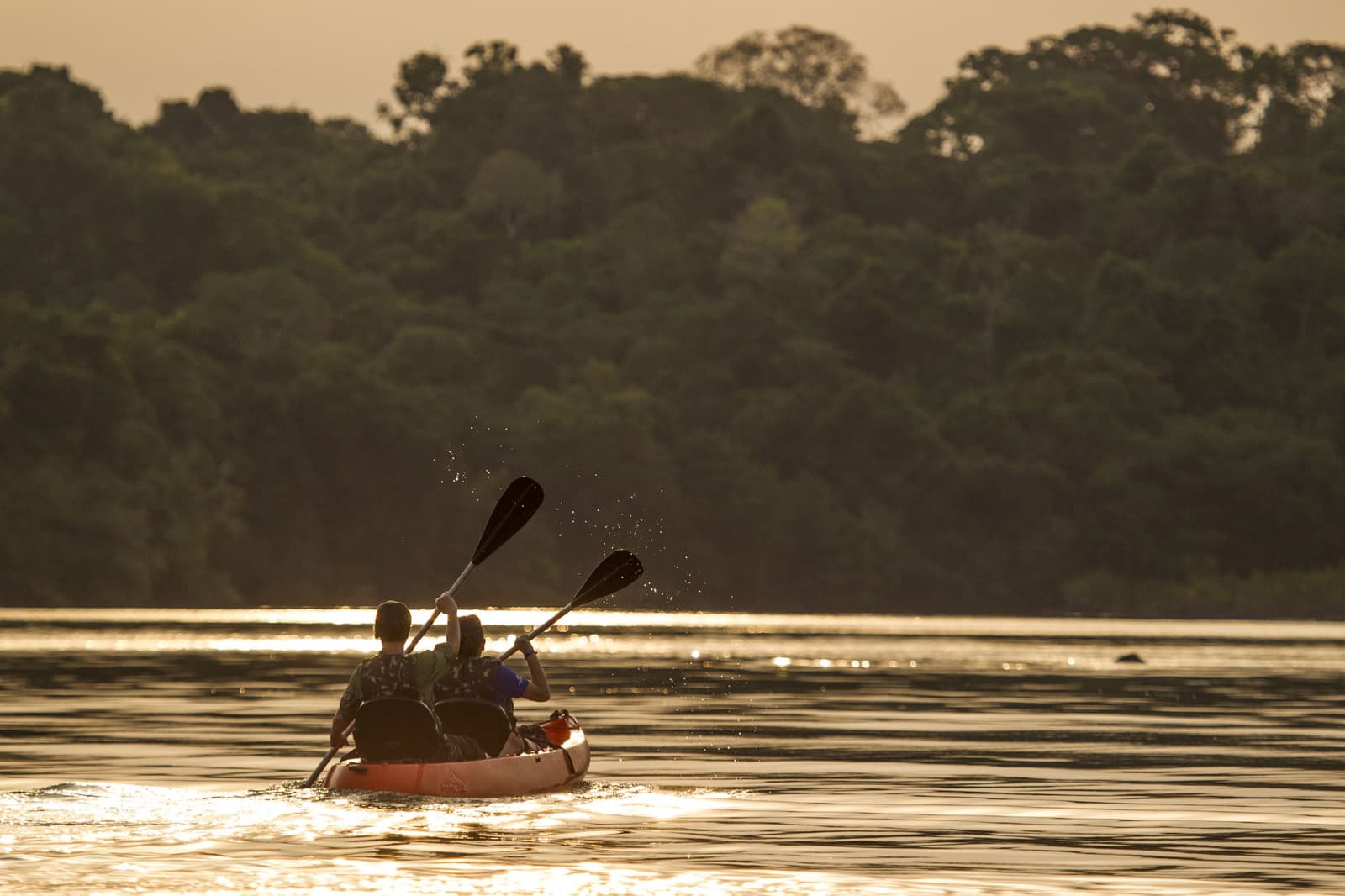 Der Fluss gibt das Tempo vor, und das ist genau richtig. Zwei Paddler in einem Tandem-Kajak gleiten bei goldenem Abendlicht über einen spiegelglatten Fluss, umgeben von dichtem Regenwalddach am Cristalino Jungle Lodge
