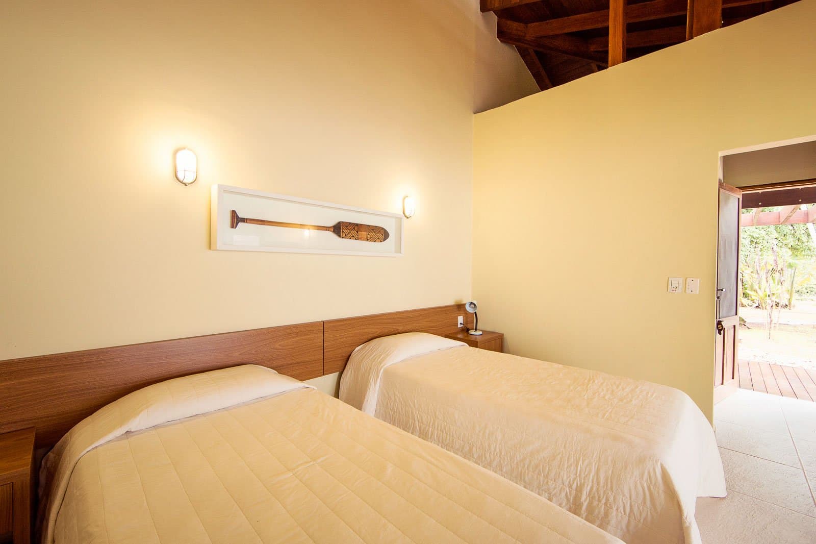 Ruhig, warm und ein Paddel überm Bett, der Amazonas bringt Stil ins Zimmer. Zweibettzimmer im Cristalino Jungle Lodge mit warmem Holzkopfteil, cremefarbener Bettwäsche, dekorativem Paddel an der gelben Wand, freiliegenden Holzbalken und offenem Gartenausgang