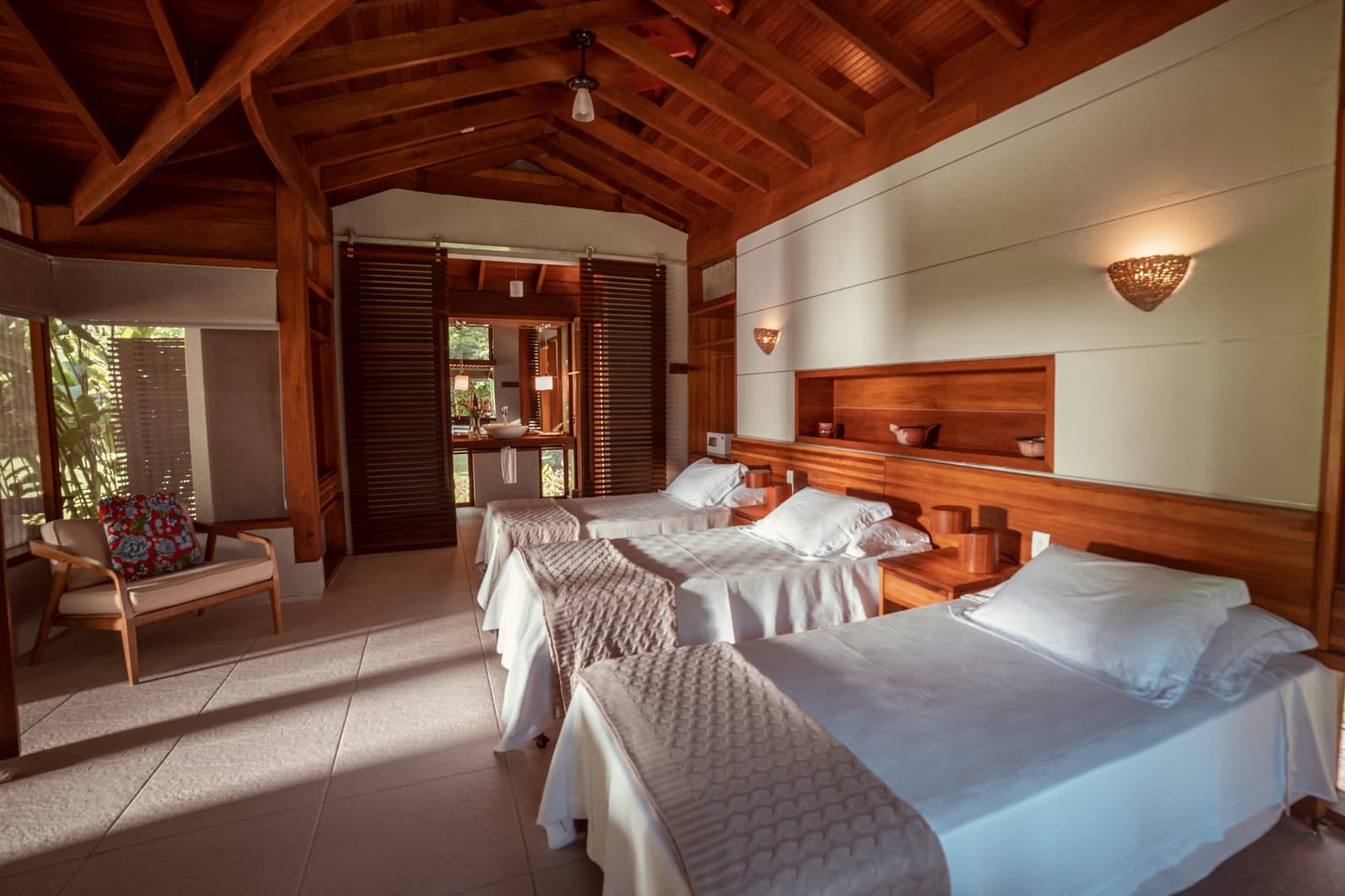 Edles Holz, frische Bettwäsche, und draußen wartet der Regenwald. Zweibettzimmer im Cristalino Jungle Lodge mit gewölbter Holzdecke, warmem Wandlicht, Holzkopfteil und Blick auf den tropischen Garten durch Lamellentüren