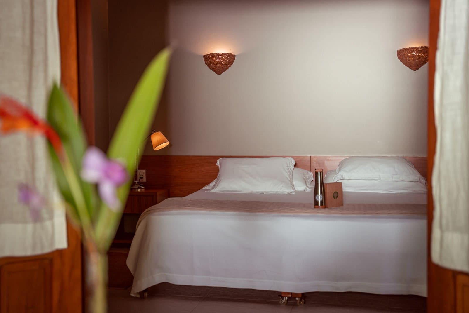 Im Amazonas schläft man überraschend tief und fest. Ein sauber gemachtes Doppelbett mit weißer Bettwäsche und warmen Holzwandleuchten in einem Gästezimmer der Cristalino Jungle Lodge, mit einer unscharfen Tropikblume im Vordergrund