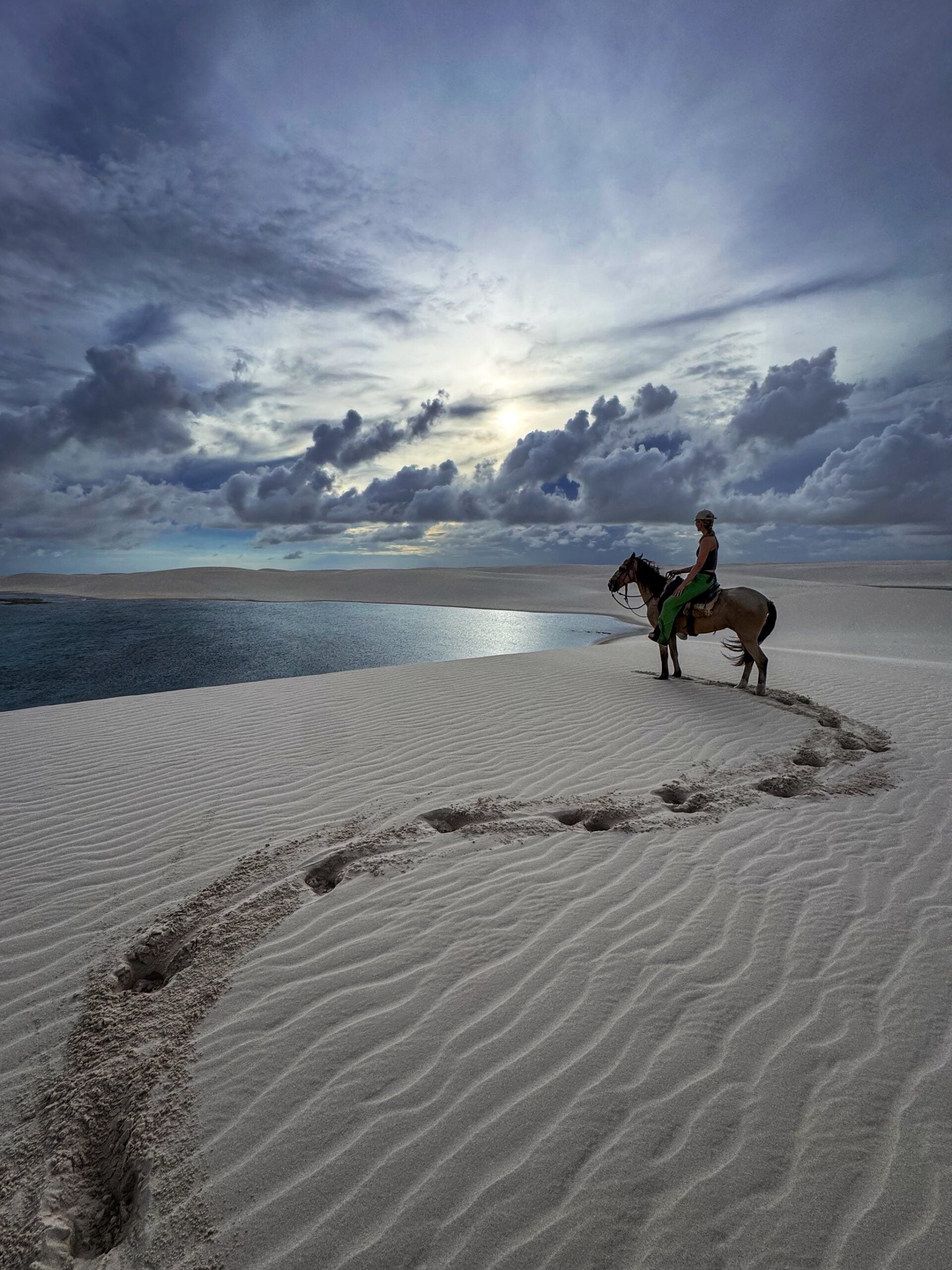 A solo rider crosses the white sand dunes of Lençóis Maranhenses National Park on horseback, with a rainwater lagoon stretching behind her. A solo rider crosses the white sand dunes of Lençóis Maranhenses National Park on horseback, with a rainwater lagoon stretching behind her.