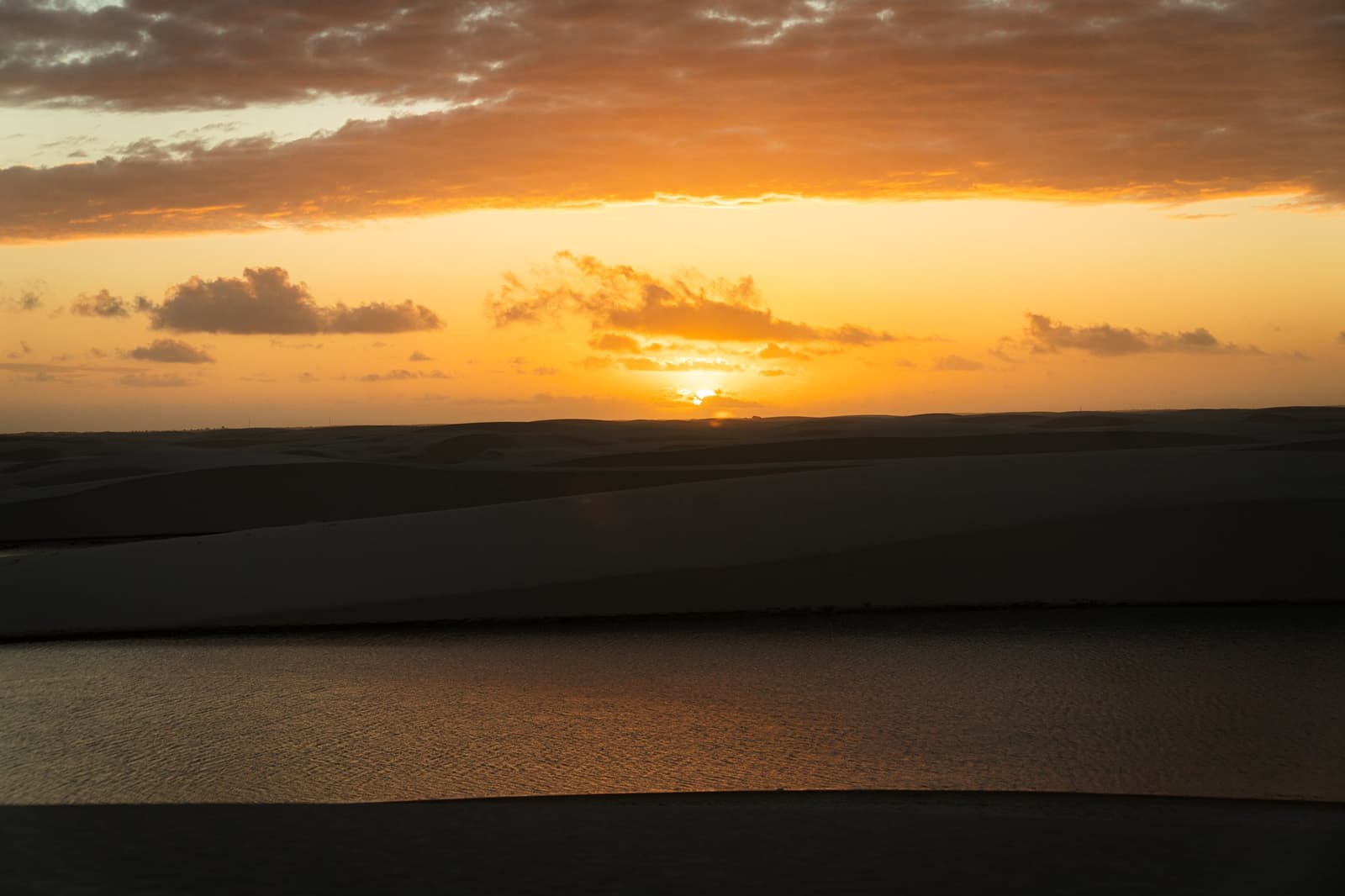 The dunes hold onto the light a little longer than everywhere else. Golden sunset over silhouetted sand dunes and a still lagoon reflecting amber light on the horseback riding Lençóis Maranhenses tour