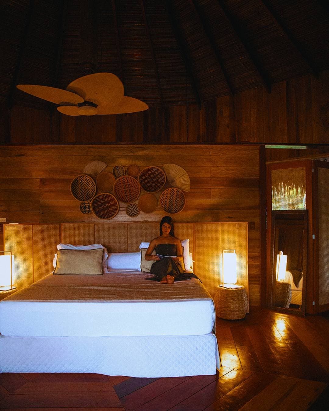 Ein gutes Buch, warmes Licht, der Abend braucht nichts weiter. Eine Frau liest auf einem Kingsize-Bett im Bungalow der Juma Amazon Jungle Lodge – warmes Lampenlicht, ein geflochtenes Rattankopfteil, dekorative Körbe an Holzwänden und eine Reetdecke mit Holzventilator