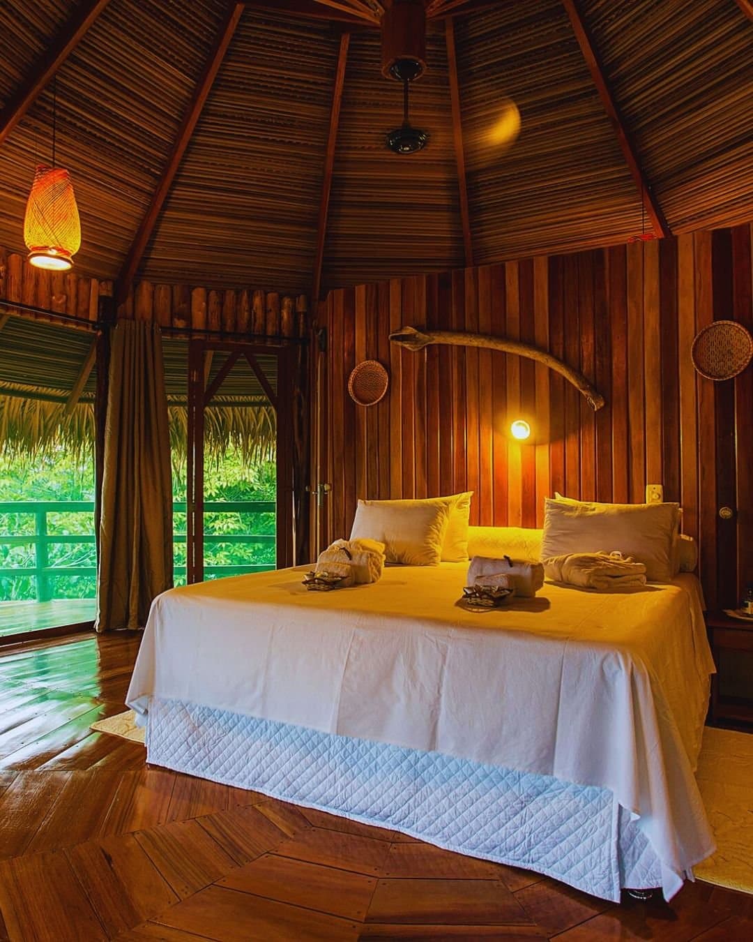 Einschlafen im Wald, aufwachen mittendrin. Warmes Bungalow-Interieur der Juma Amazon Jungle Lodge mit Doppelbett in weißer Bettwäsche, kegelförmigem Strohdach, poliertem Hartholzboden und einem offenen Balkon mit Blick auf üppige Regenwaldvegetation