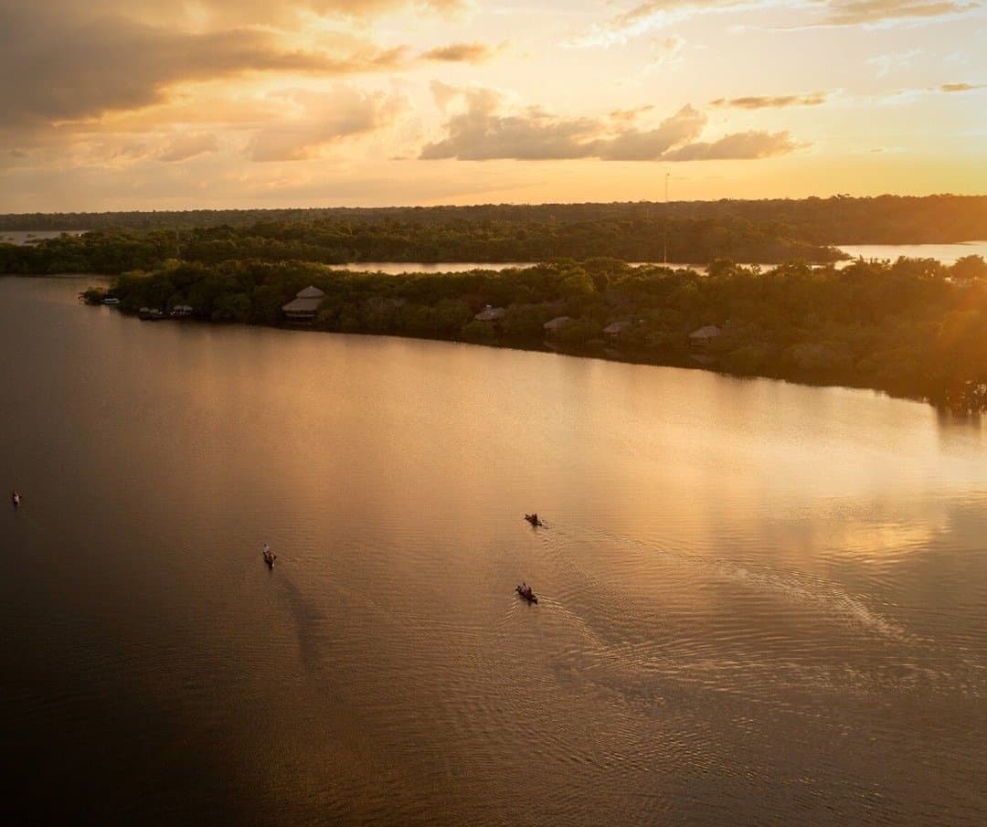Am Amazonas bestimmt der Fluss das Tempo, nicht umgekehrt. Luftaufnahme kleiner Kanus auf einem breiten Fluss im goldenen Sonnenuntergangslicht, mit den Strohdach-Bungalows der Juma Amazon Jungle Lodge im dichten Regenwald am gegenüberliegenden Ufer