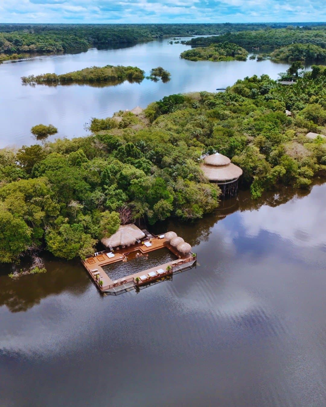 Ein eigener Flussabschnitt, mitten im Blätterdach. Luftaufnahme der Juma Amazon Jungle Lodge mit einem schwimmenden Holzdeck, strohgedeckten Pavillons und einem Flusspool inmitten überschwemmten Regenwaldes