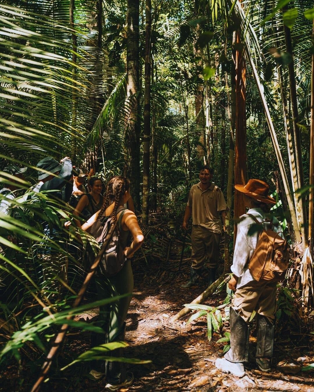 Der Wald hat seinen eigenen Takt, und ein guter Guide hilft, ihn zu spüren. Eine kleine Gruppe lächelnder Reisender folgt einem einheimischen Führer auf einem schmalen Pfad durch dichten tropischen Regenwald der Juma Amazon Jungle Lodge, umgeben von Palmwedeln und hohen Bäumen