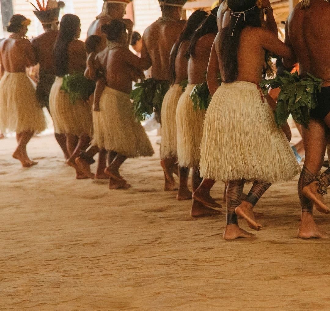 Jeder Schritt sitzt. Kultur, die lebt, nicht inszeniert. Eine Reihe indigener Tänzerinnen und Tänzer in geflochtenen Strohröcken und Blattschmuck bewegt sich barfuß über einen Sandboden bei einer traditionellen Zeremonie während des Gemeinschaftsbesuchs in der Juma Amazon Jungle Lodge