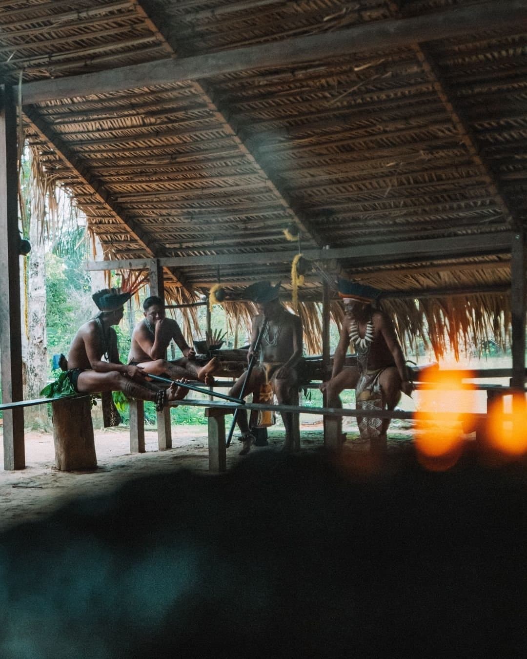 In der Maloca zählt das Gespräch mehr als die Uhrzeit. Mitglieder einer indigenen Gemeinschaft in traditioneller Körperbemalung, Kopfschmuck und Perlenketten versammeln sich unter einem strohgedeckten Freiluftgebäude der Juma Amazon Jungle Lodge, lange Blasrohre lehnen auf Holzbänken