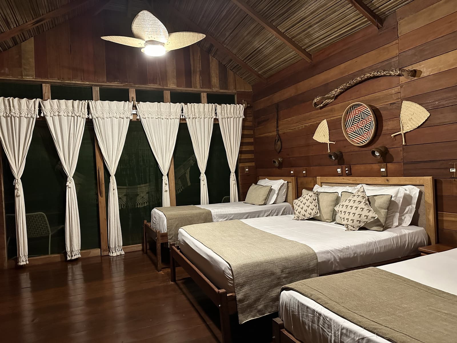 Handwerk an den Wänden, frisches Leinen auf den Betten, so schläft man gut im Amazonas. Geräumiges Bungalow der Juma Amazon Jungle Lodge bei Nacht mit drei Holzbetten in weißer Bettwäsche, einem geflochtenen Deckenventilator und einheimischen Korbwaren und Paddeln an den Holzwänden