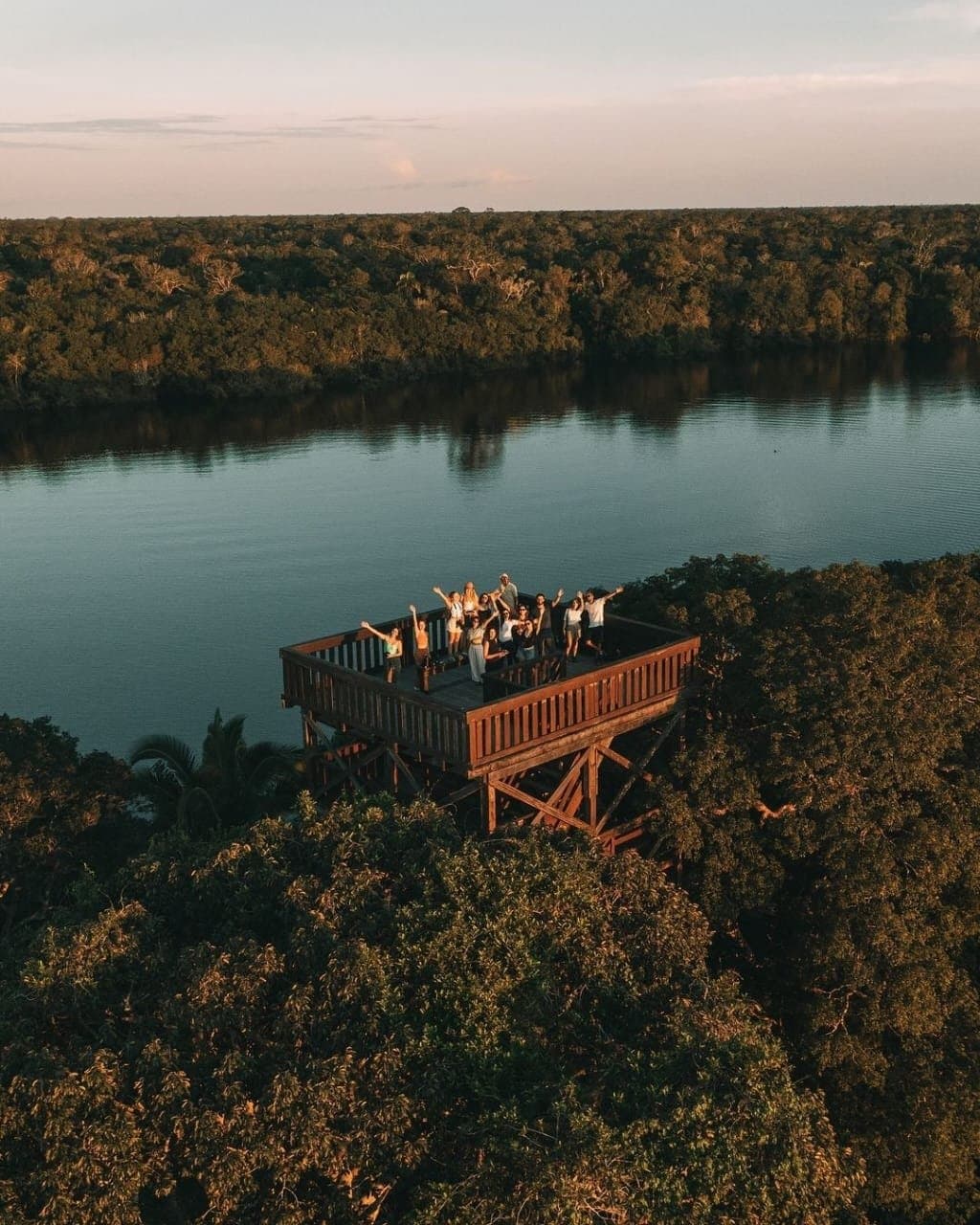 Arme hoch, Blätterdach unten, Fluss voraus, so verabschiedet sich der Amazonas vom Tag. Eine Reisegruppe winkt begeistert von einem hohen hölzernen Aussichtsturm der Juma Amazon Jungle Lodge, umgeben von dichtem Regenwalddach und einem ruhigen Fluss im Sonnenuntergang
